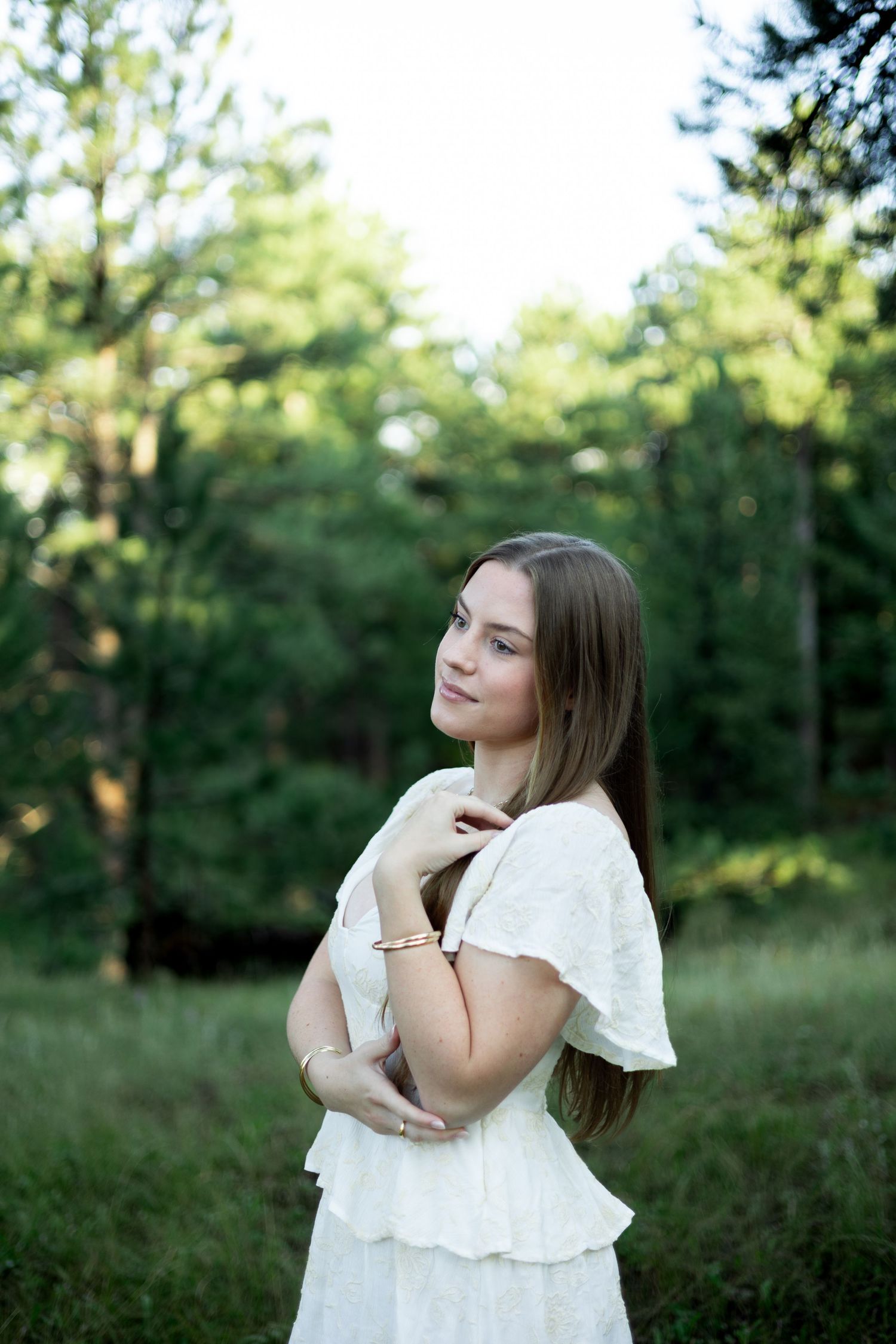 A person in a white ruffled dress poses thoughtfully in a sunlit outdoor setting with green foliage in the background.