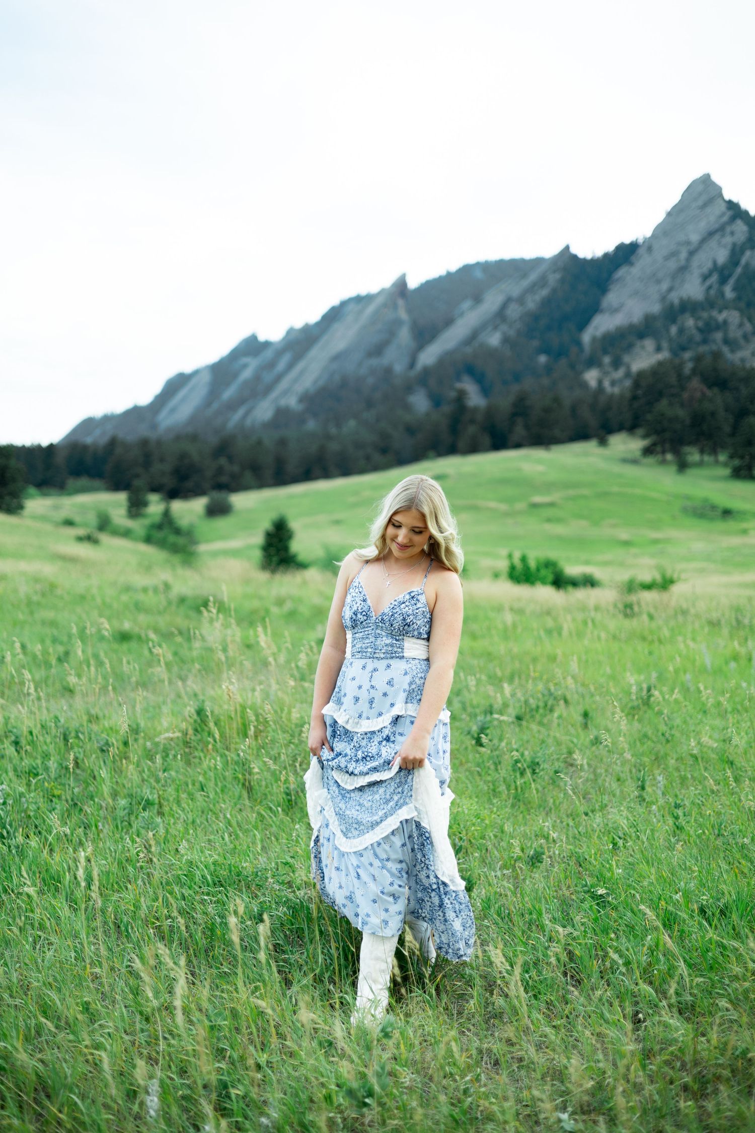 Woman in blue floral dress walks through green mountain meadow with dramatic peaks rising in background.