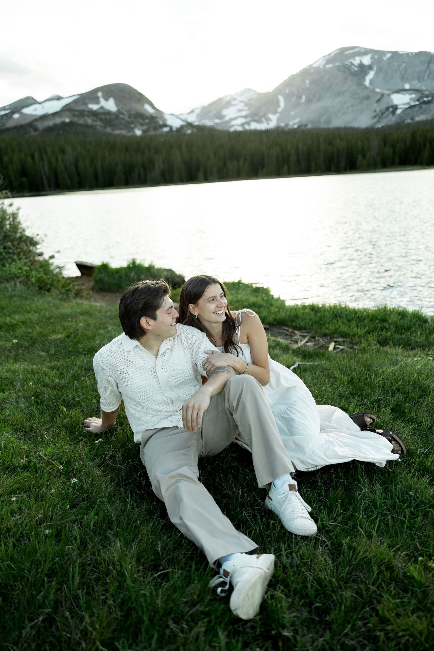 A couple in wedding attire sits on green grass by a mountain lake with snow-capped peaks and pine trees in the background.