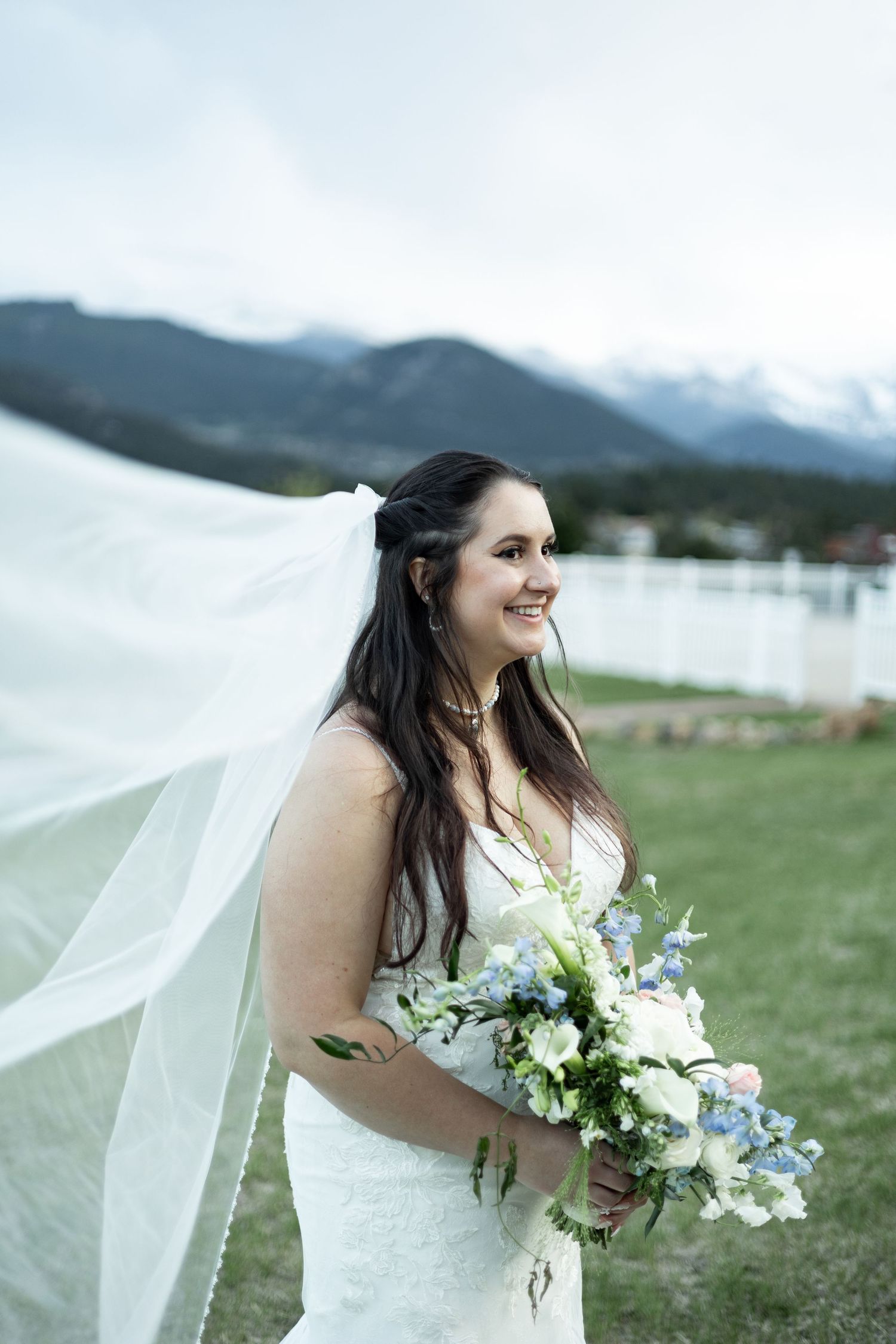 A bride in a white lace dress holds a blue and white bouquet as her veil flows in the wind near mountains and a lake.