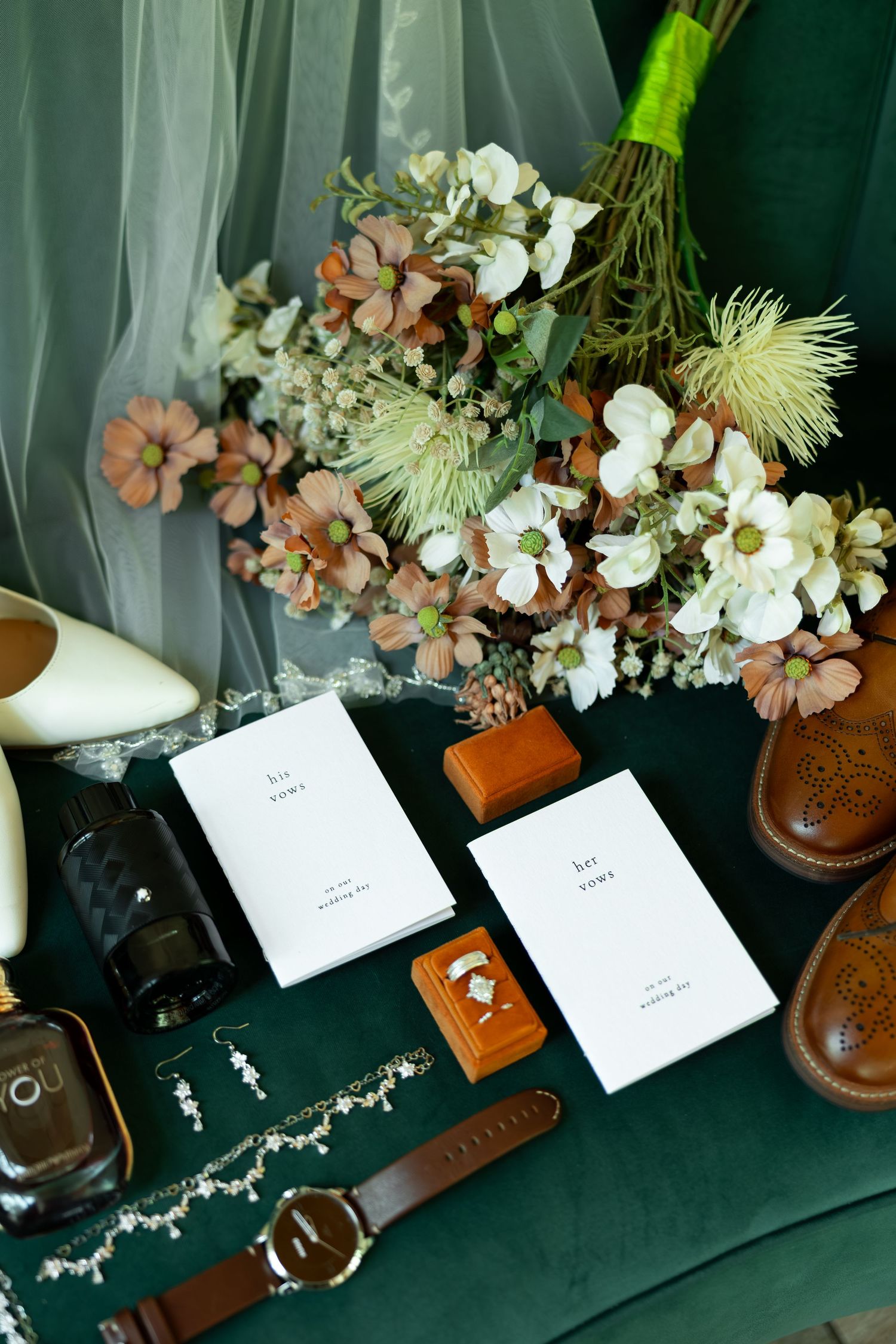 Wedding flat lay with vow books, rings, bridal bouquet, heels, and accessories on green velvet surface.