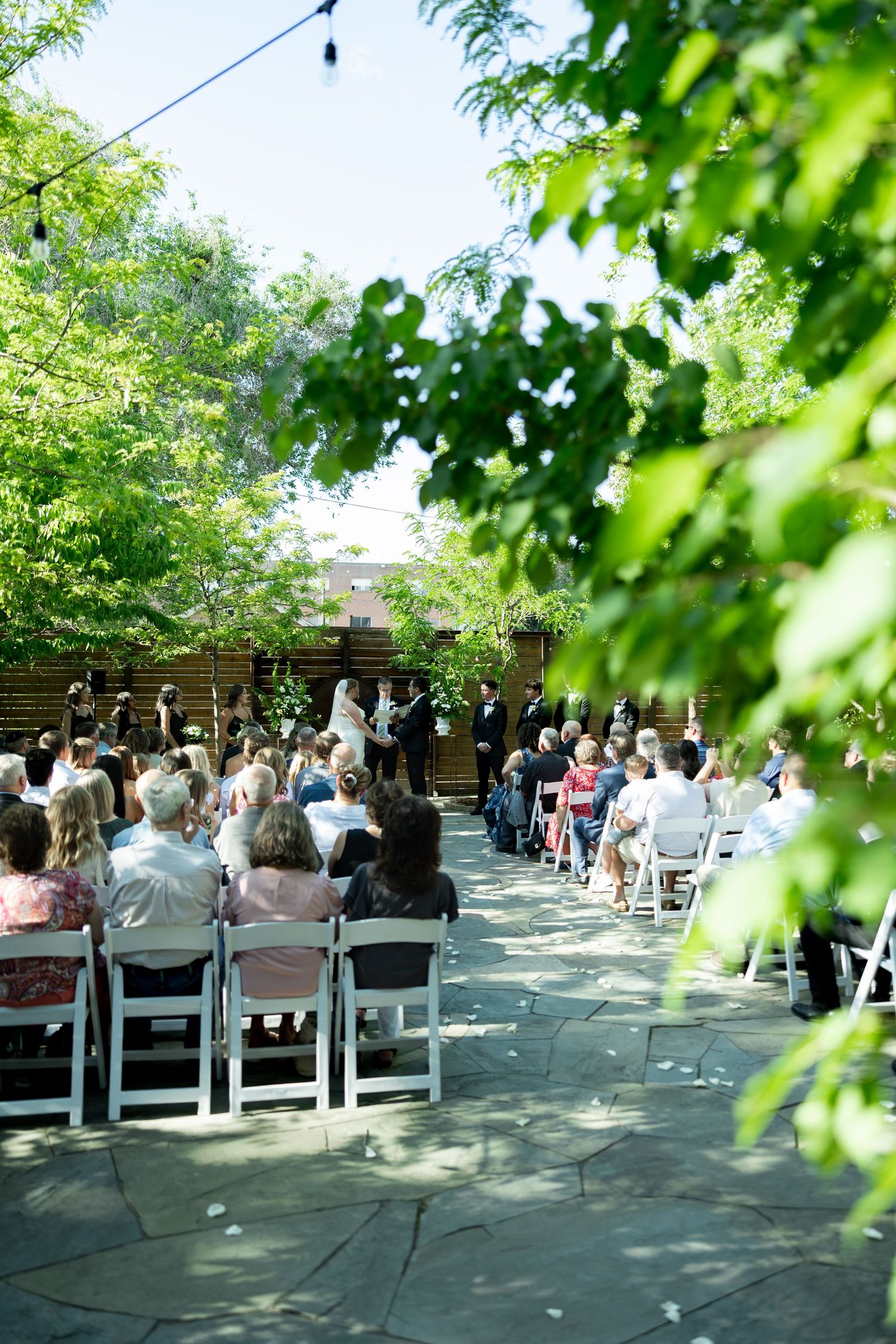 Outdoor wedding ceremony with guests seated in white chairs along a stone path surrounded by lush green trees.