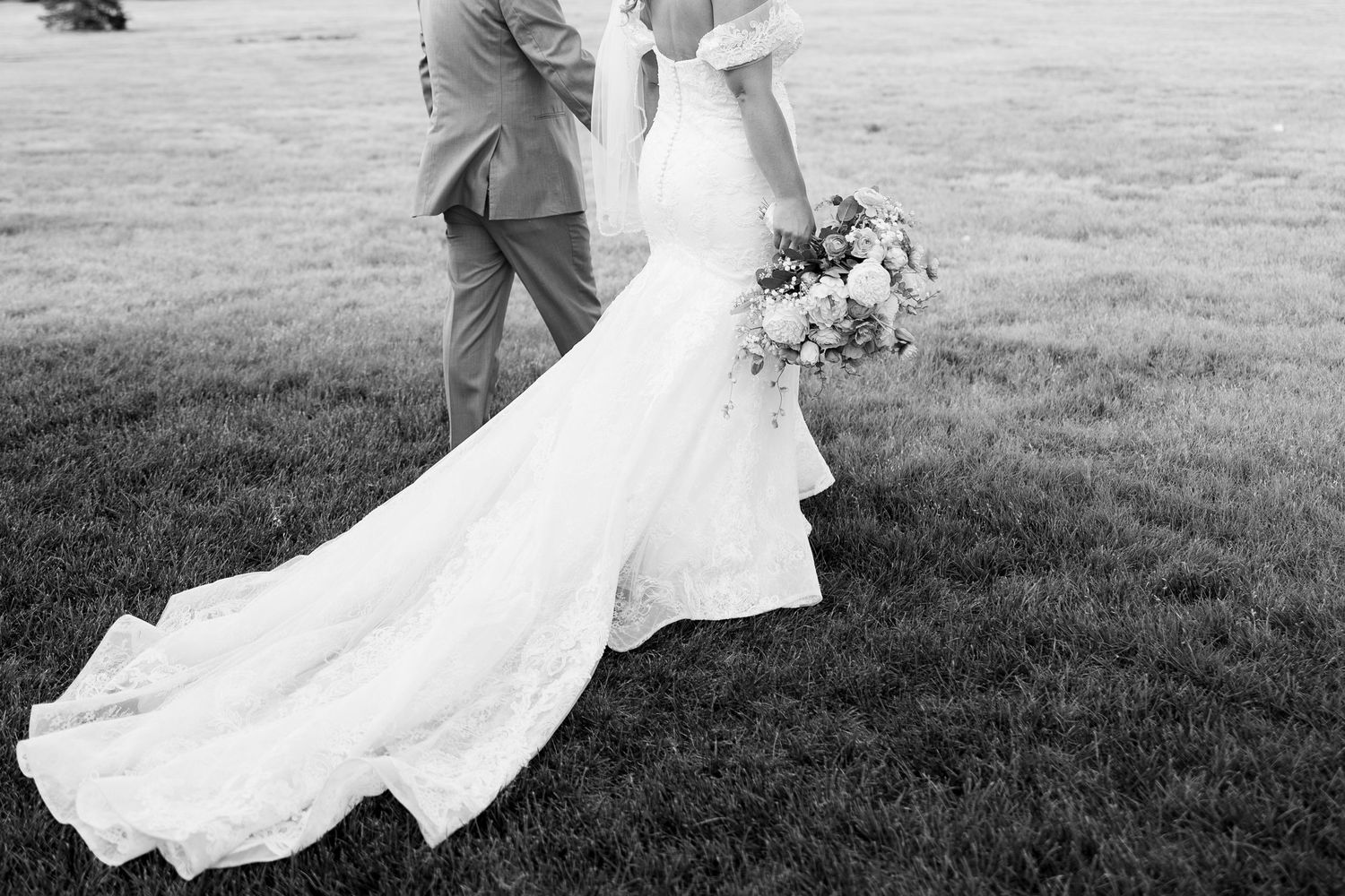 Black and white photo of bride in lace gown with train holding bouquet beside groom on green lawn.