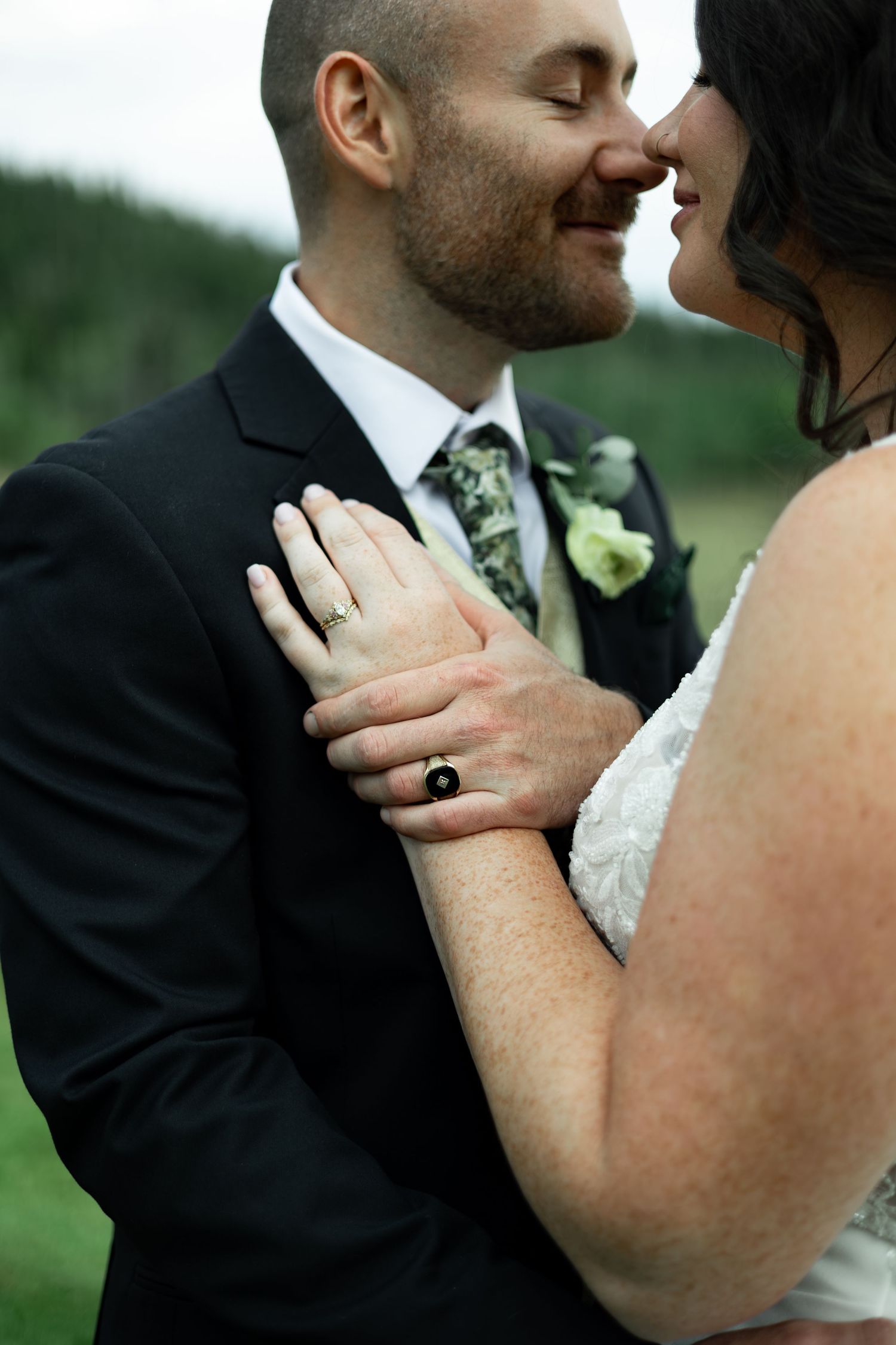 Bride and groom share a romantic moment, her hand with black ring resting on his dark suit jacket outdoors.