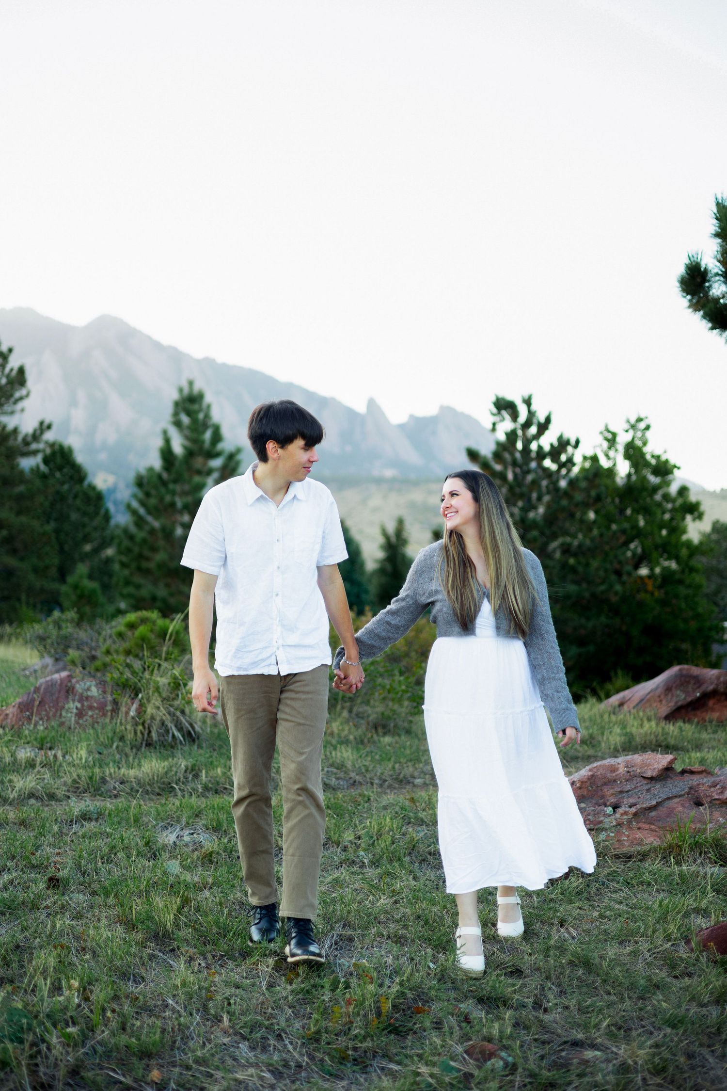 A couple holds hands while walking through a mountain meadow, smiling at each other in casual outdoor attire.