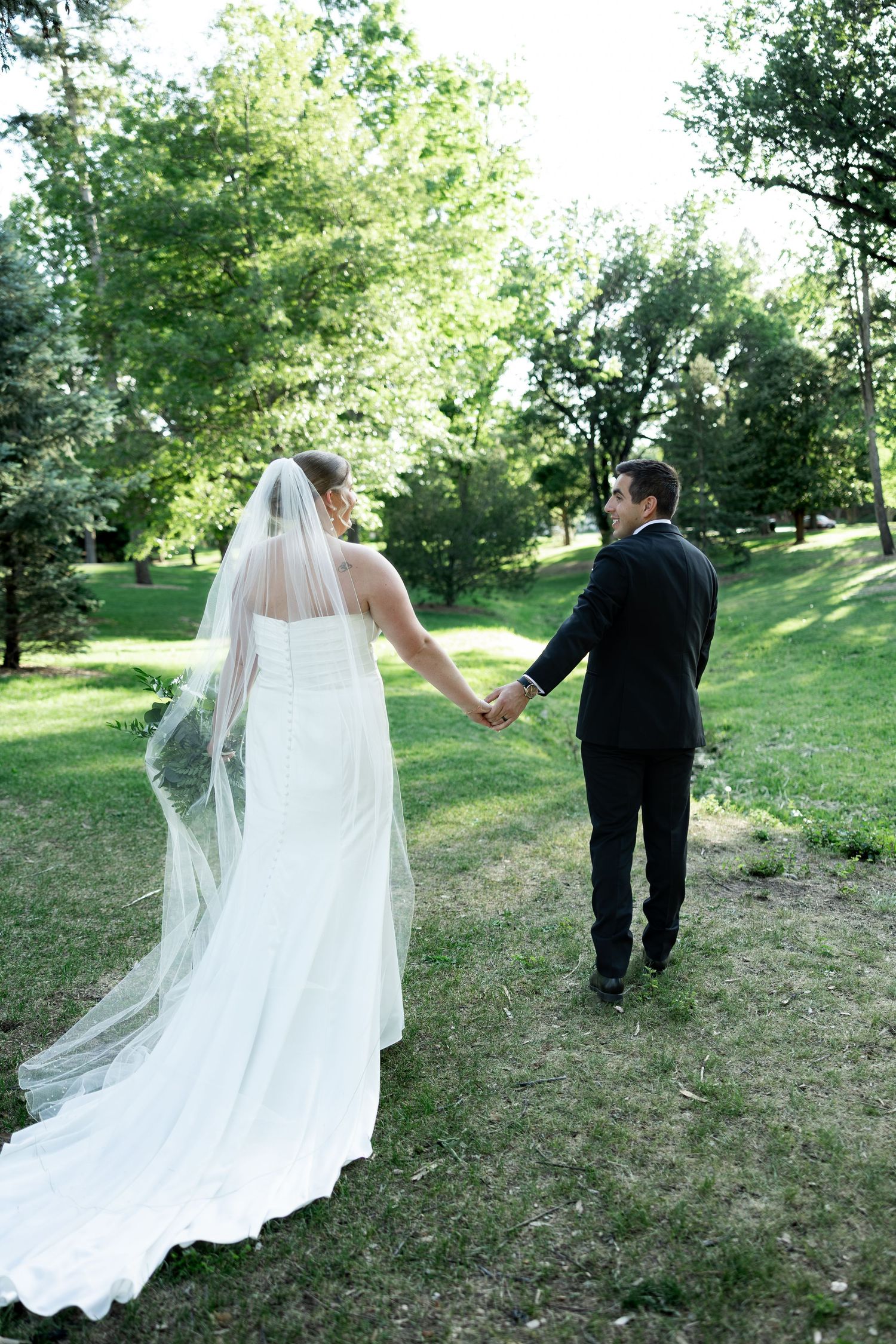 Bride in white gown and veil holds hands with groom in black suit walking through green park.