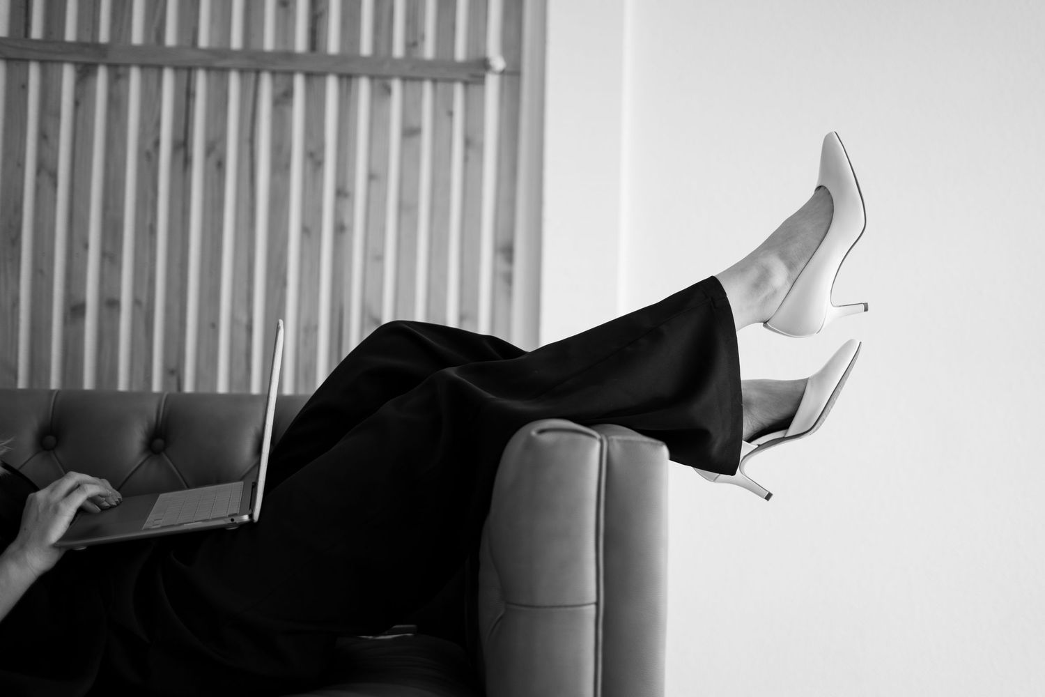Black and white photo of legs in formal black pants and white high heels resting on a modern chair.