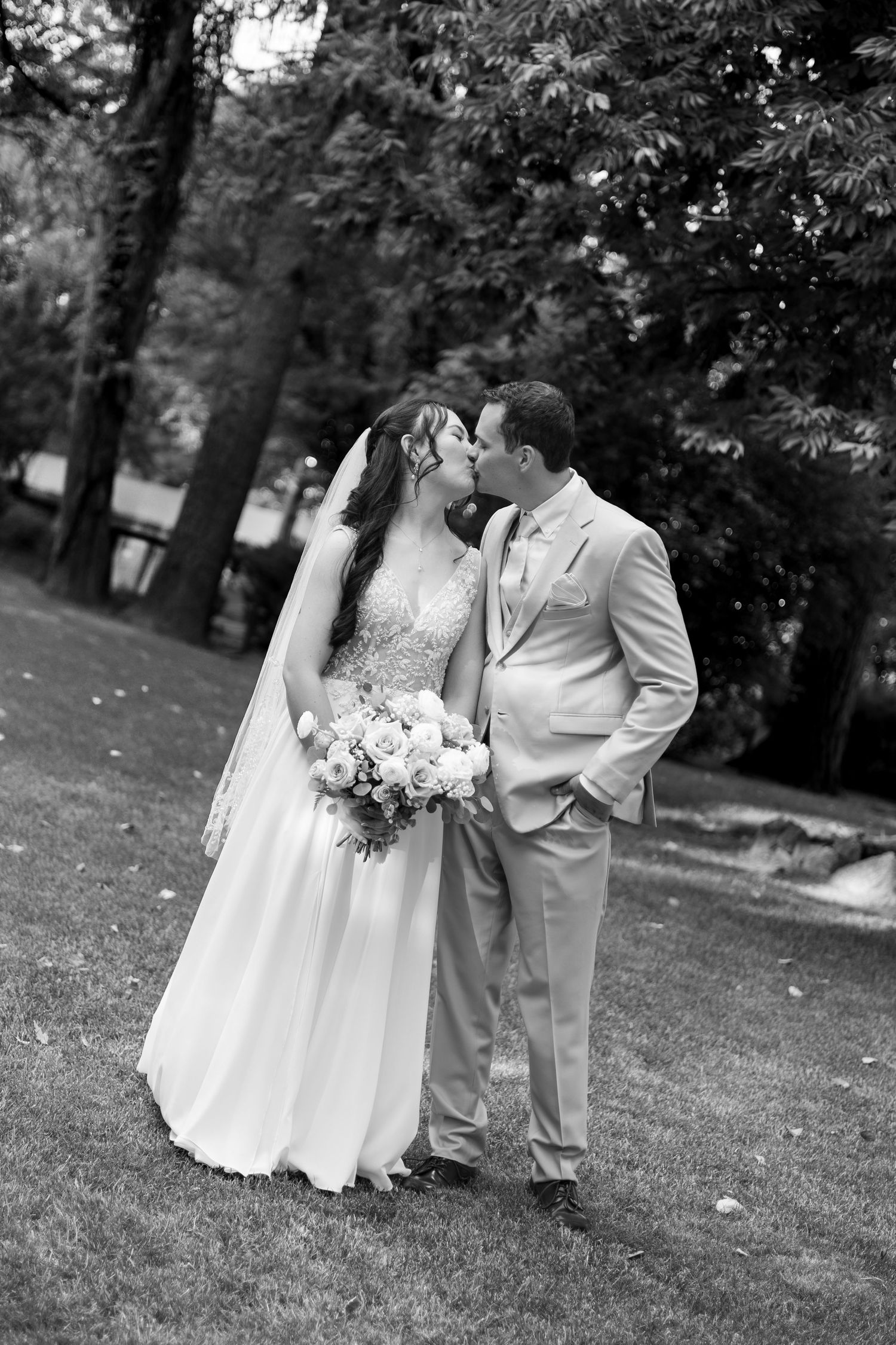 Black and white photo of a bride and groom sharing a kiss outdoors, bride holding a bouquet, surrounded by lush trees.