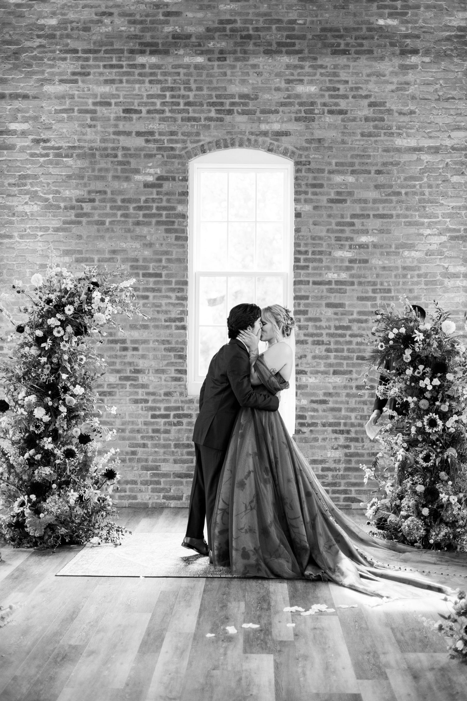 Black and white photo of a couple embracing in front of a brick wall with floral arrangements.