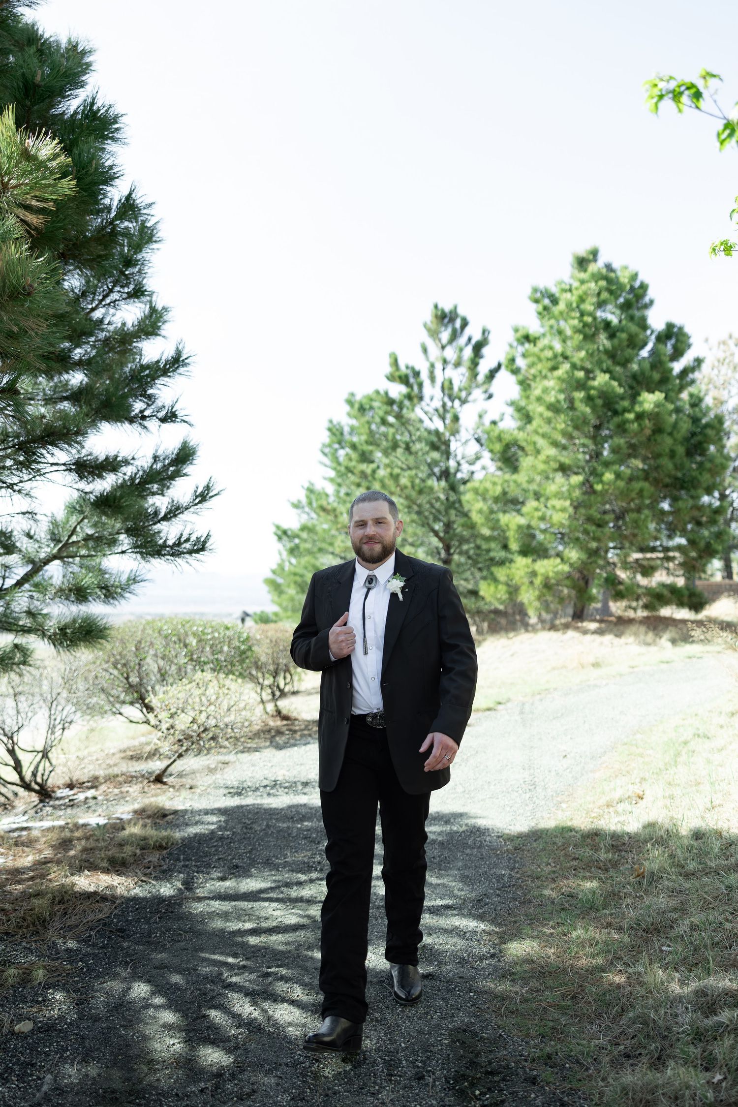 Groom in black tuxedo with white boutonniere poses on gravel path surrounded by pine trees on wedding day.