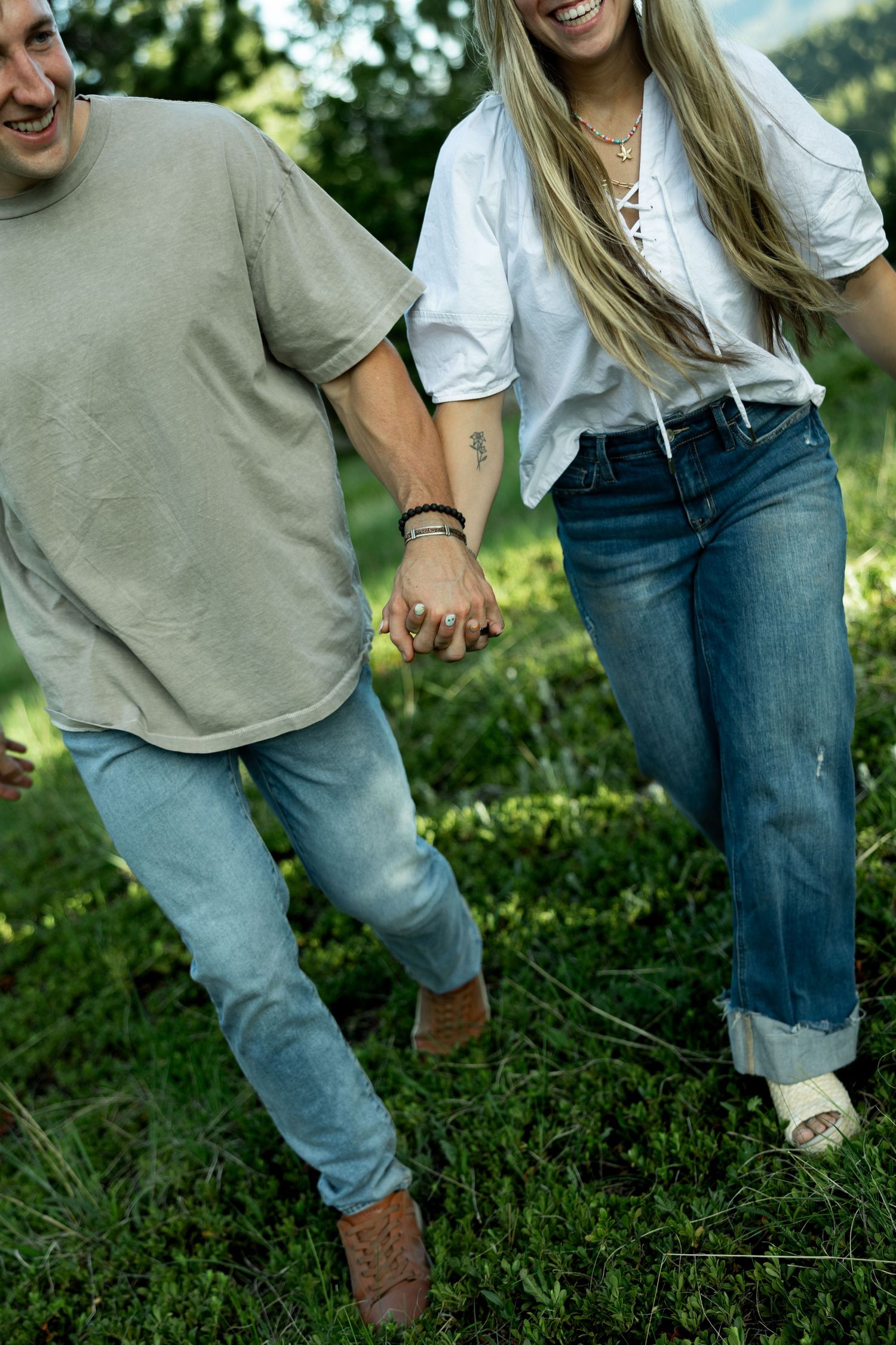 A smiling couple holds hands while walking through a lush green field, dressed casually in jeans and relaxed tops.