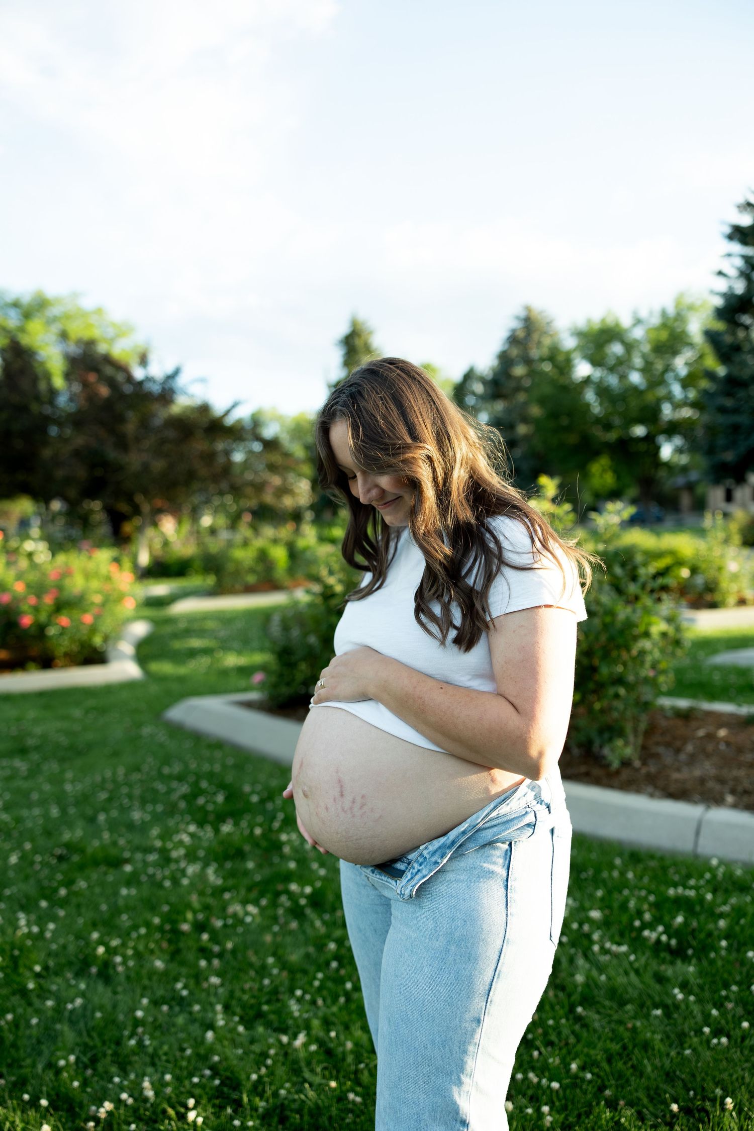 Pregnant woman in white crop top and jeans cradling baby bump outdoors in a lush green park on a sunny day.