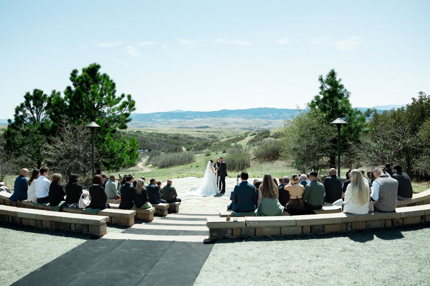 Outdoor wedding ceremony with guests seated in stone amphitheater overlooking scenic rolling hills under clear blue sky.