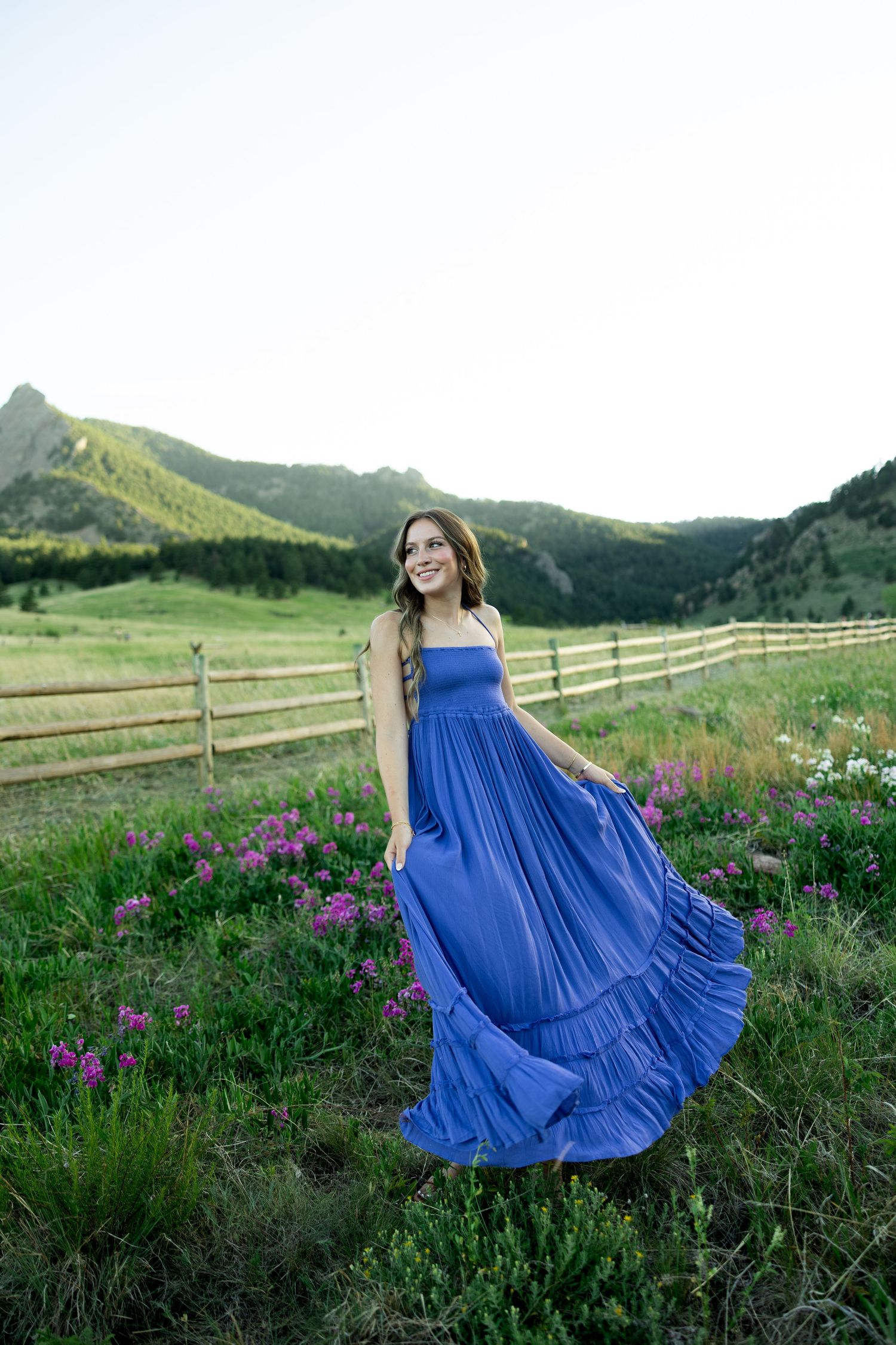 Woman in flowing blue maxi dress smiling in a mountain meadow with wildflowers and wooden fence backdrop.