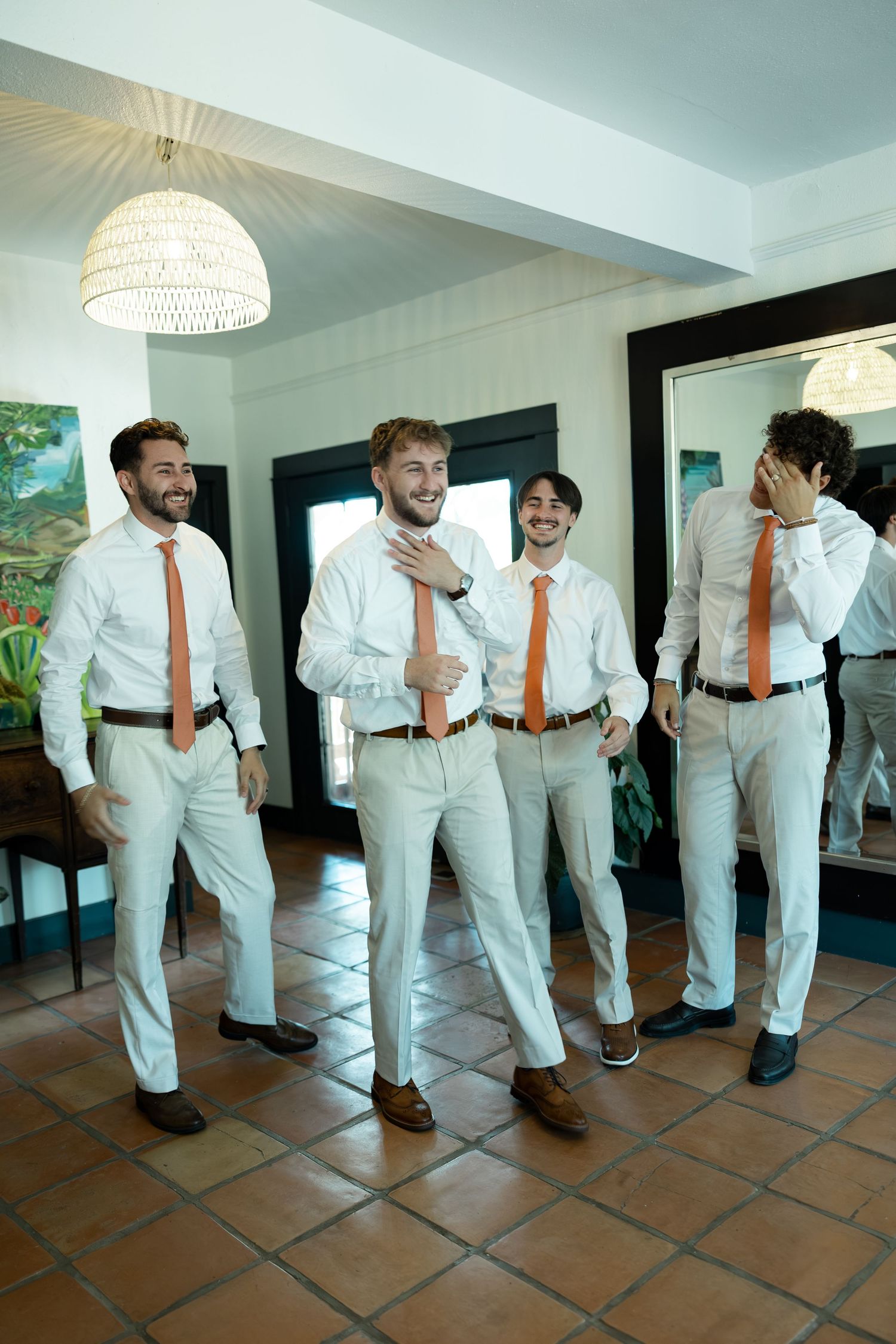 Four groomsmen in white shirts and orange ties laughing together before a wedding ceremony.