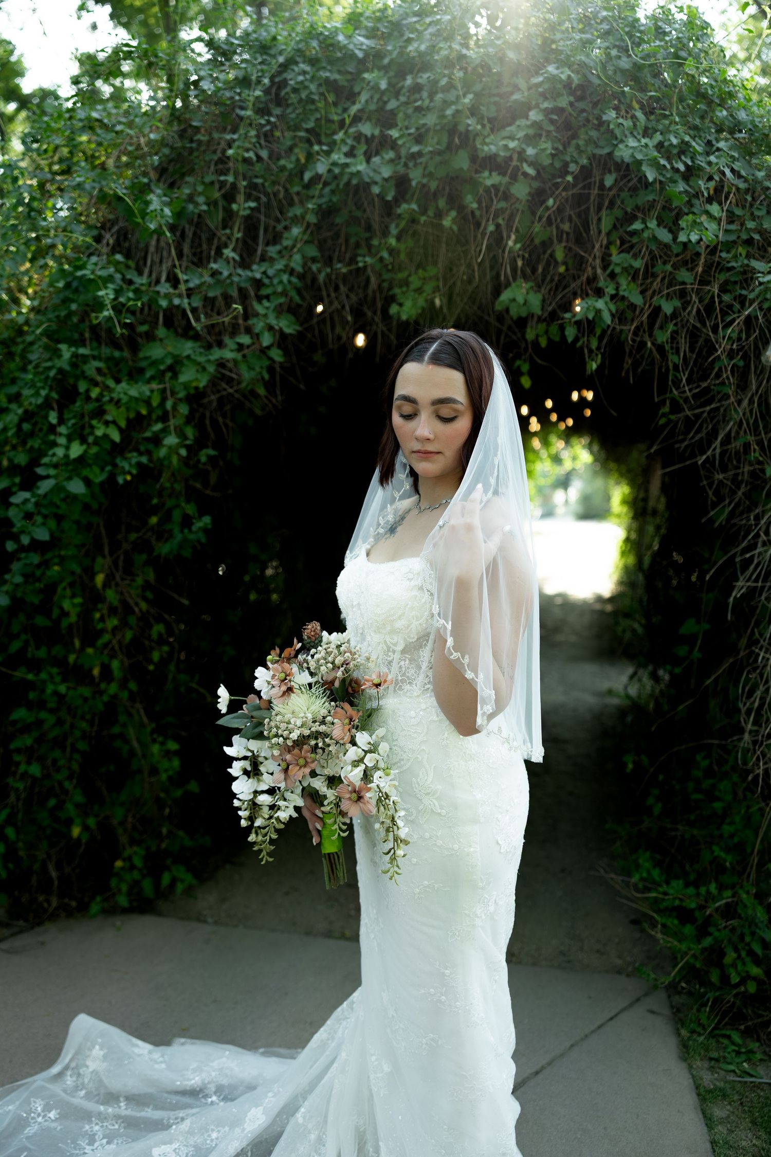 Bride in white lace gown and veil holds floral bouquet under green ivy arch outdoors.