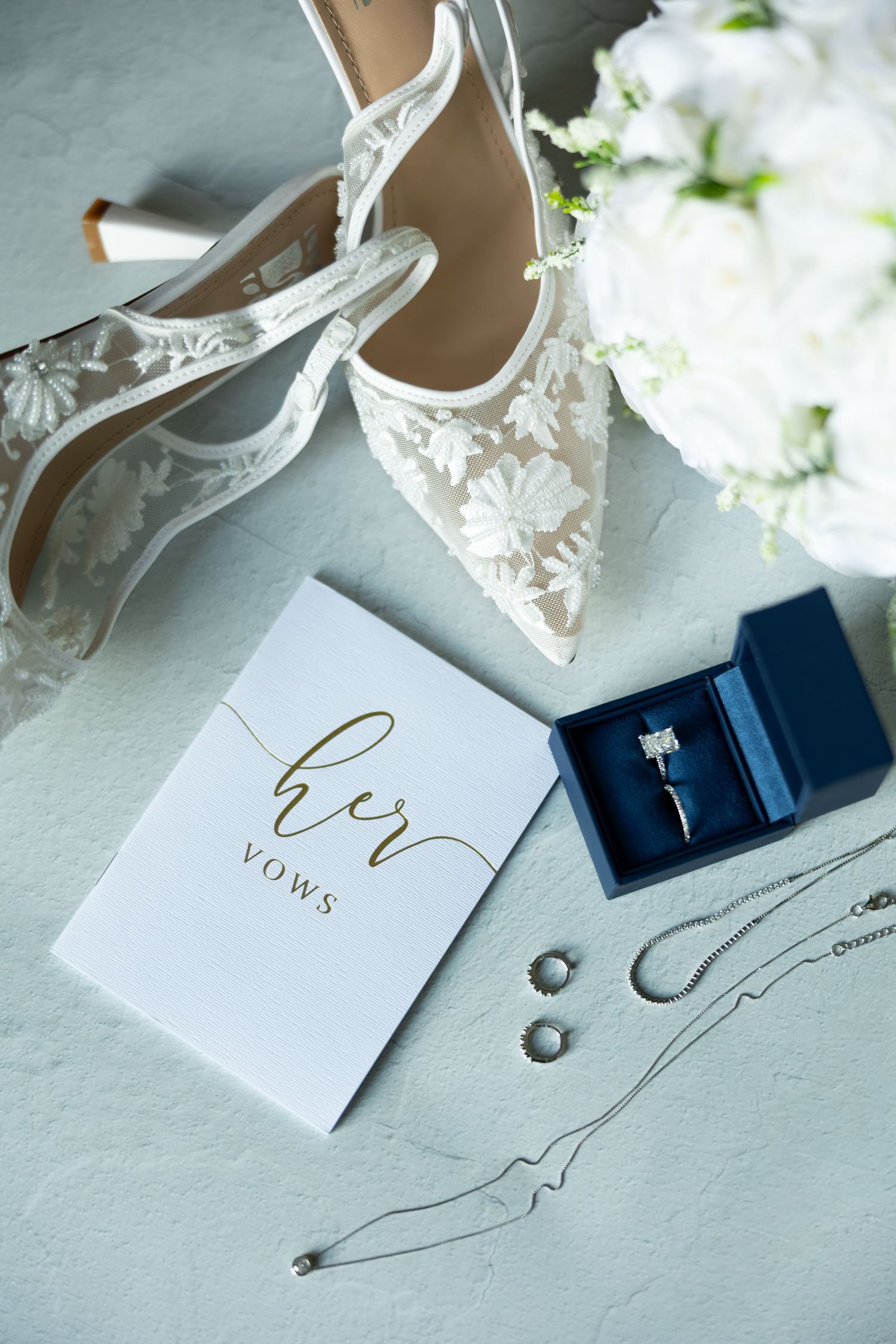 Wedding details flatlay with lace shoes, vows book, rings, and white flowers on light background.