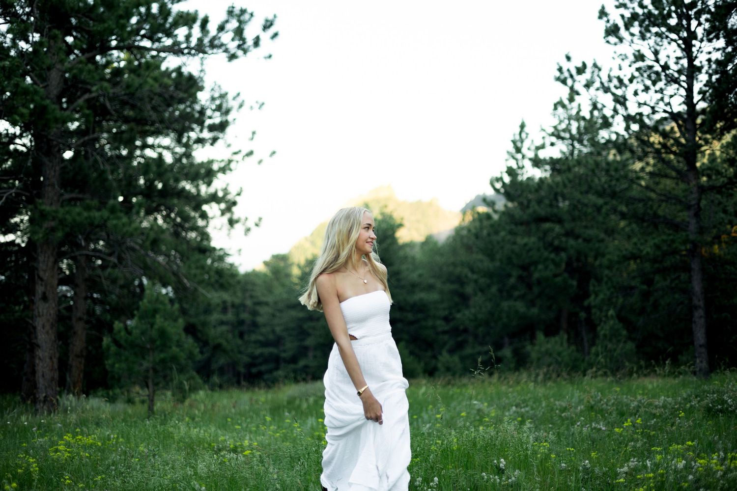 Against a backdrop of trees, someone twirls gracefully in a white dress amidst tall green grass.