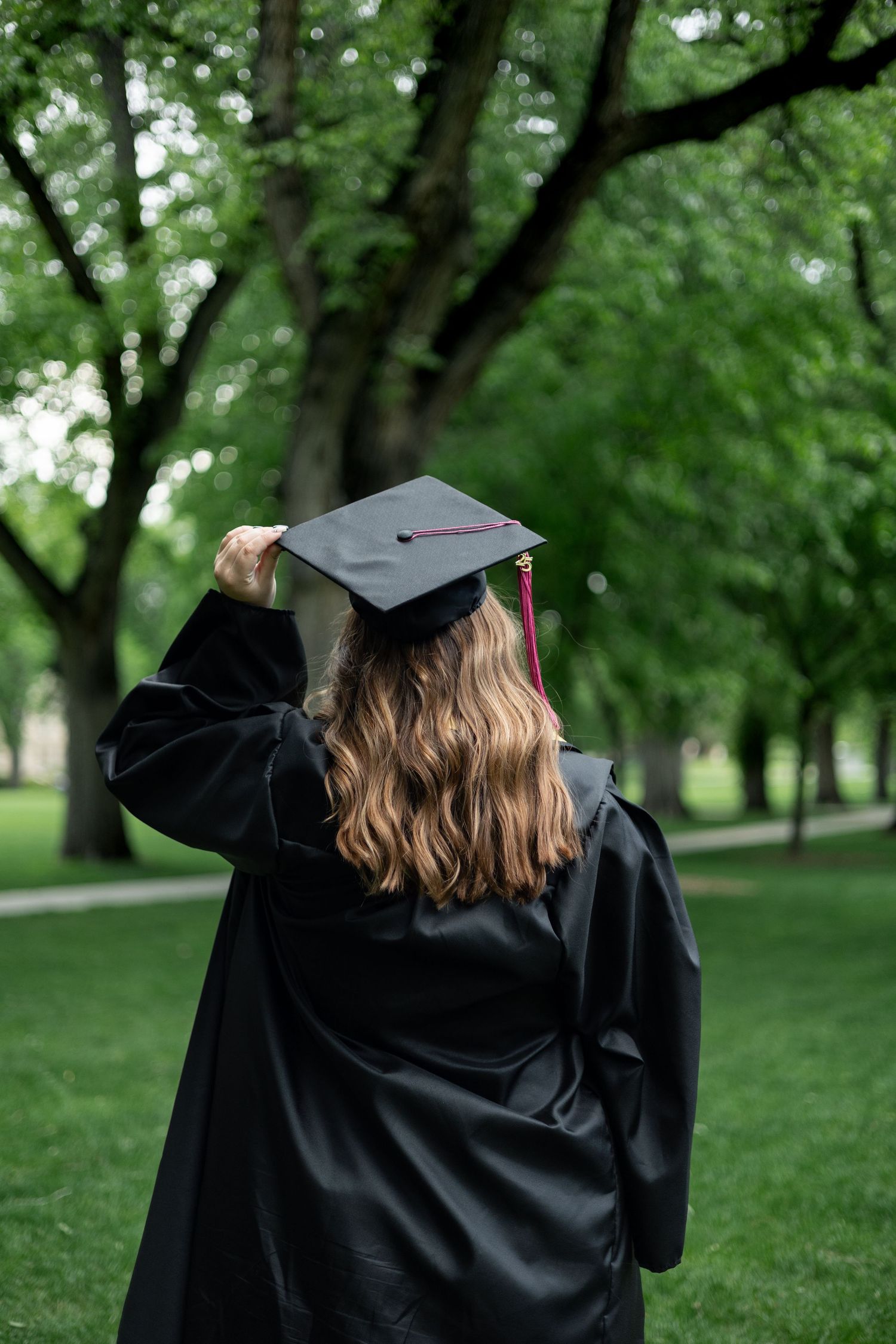 Graduate in black cap and gown touching graduation cap while standing in a lush green park setting.