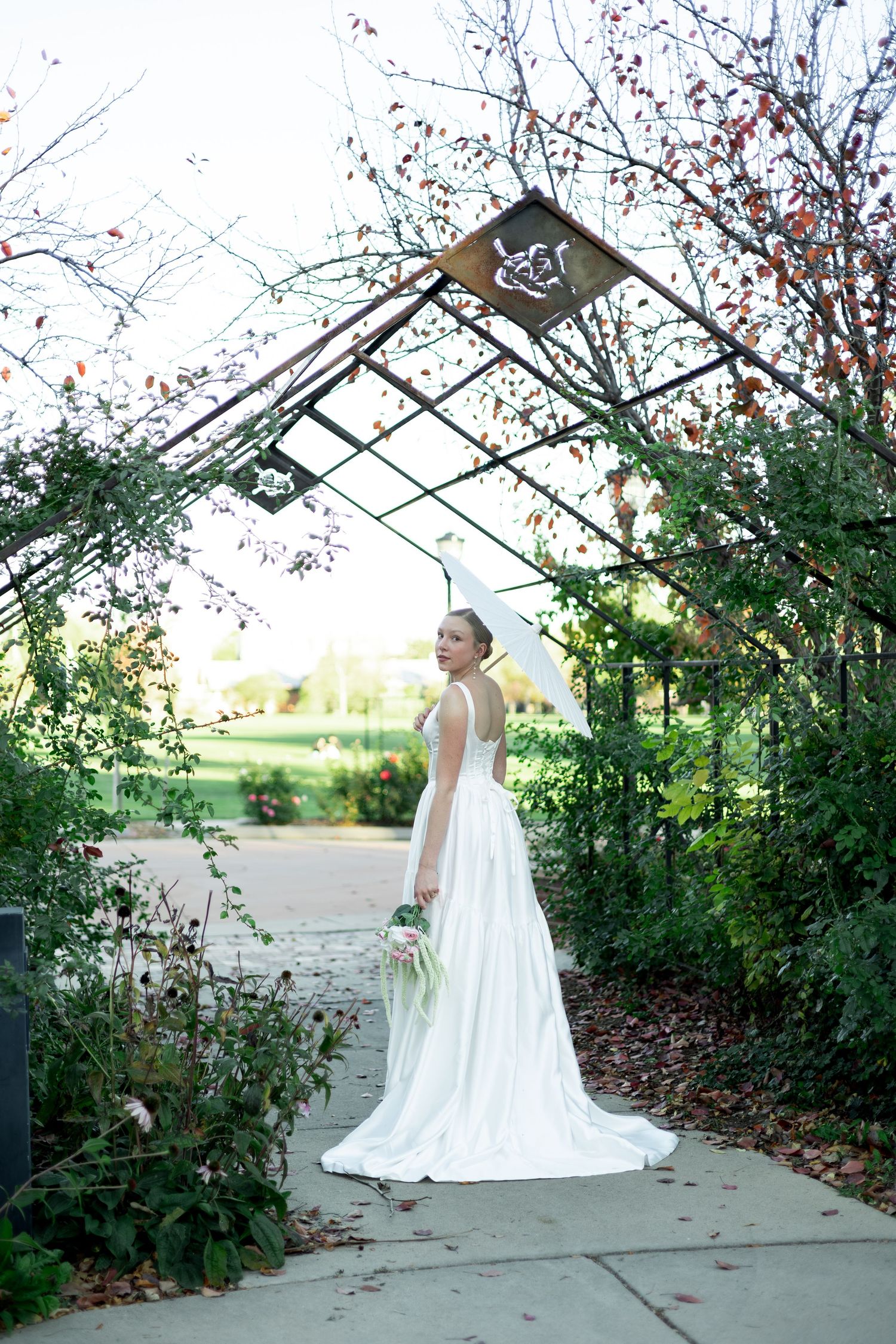 A bride in a white wedding dress poses under a garden arbor with autumn foliage and red berries at sunset.