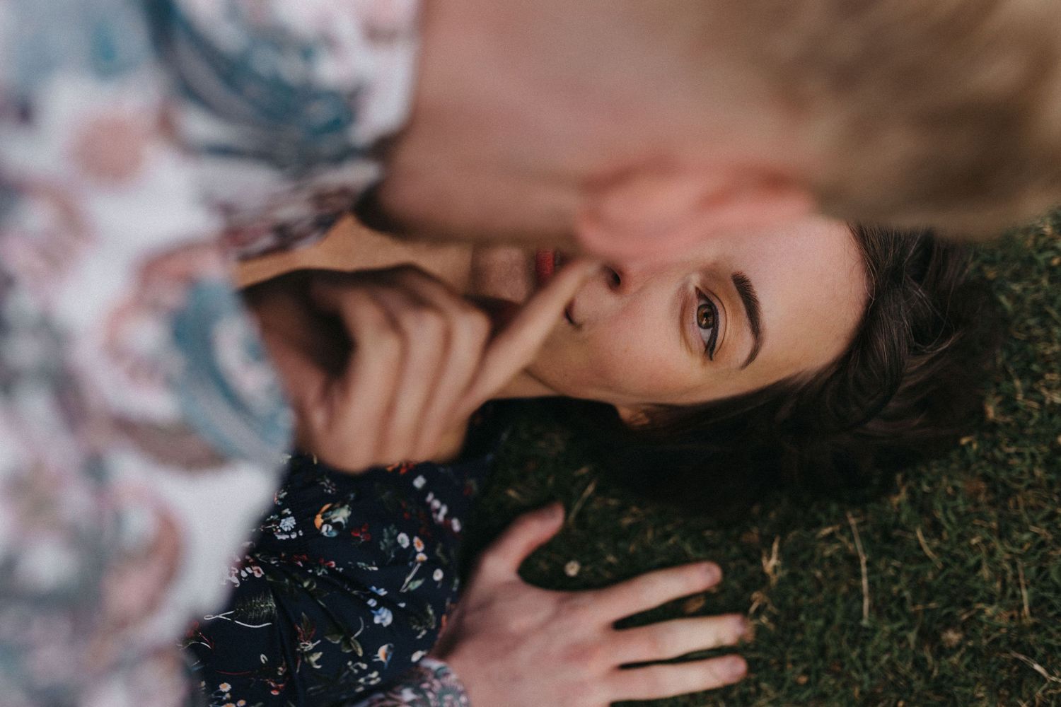 Artistic portrait of someone laying in grass with dark curly hair and dreamy makeup against a soft blurred floral backdrop.