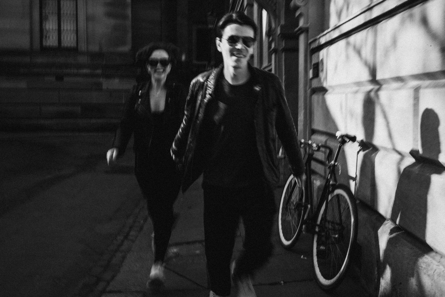 A black and white urban street scene shows people walking past a parked bicycle on a shadowy sidewalk.