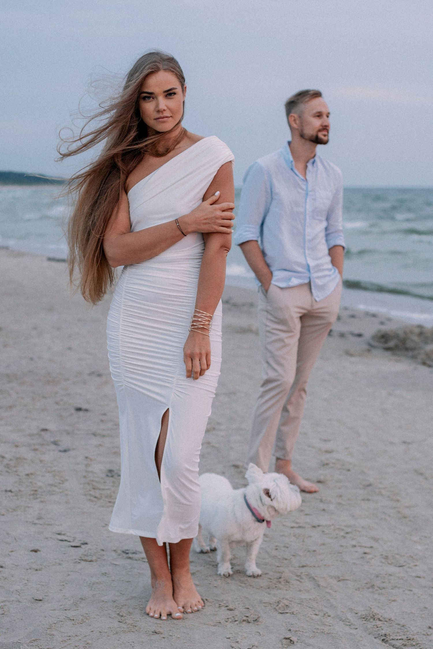 Stylish couple poses with small white dog on beach at sunset while wearing elegant white and light blue outfits.