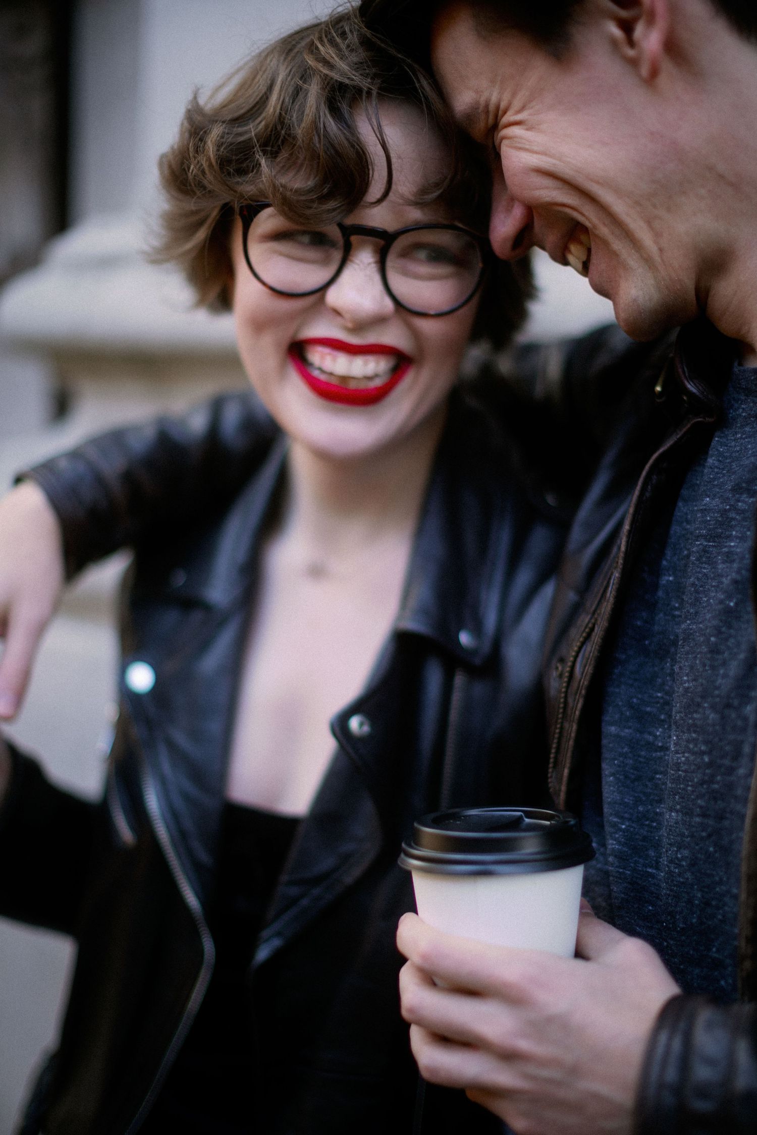 A couple shares an intimate moment while one holds a to-go coffee cup, wearing a leather jacket and glasses.