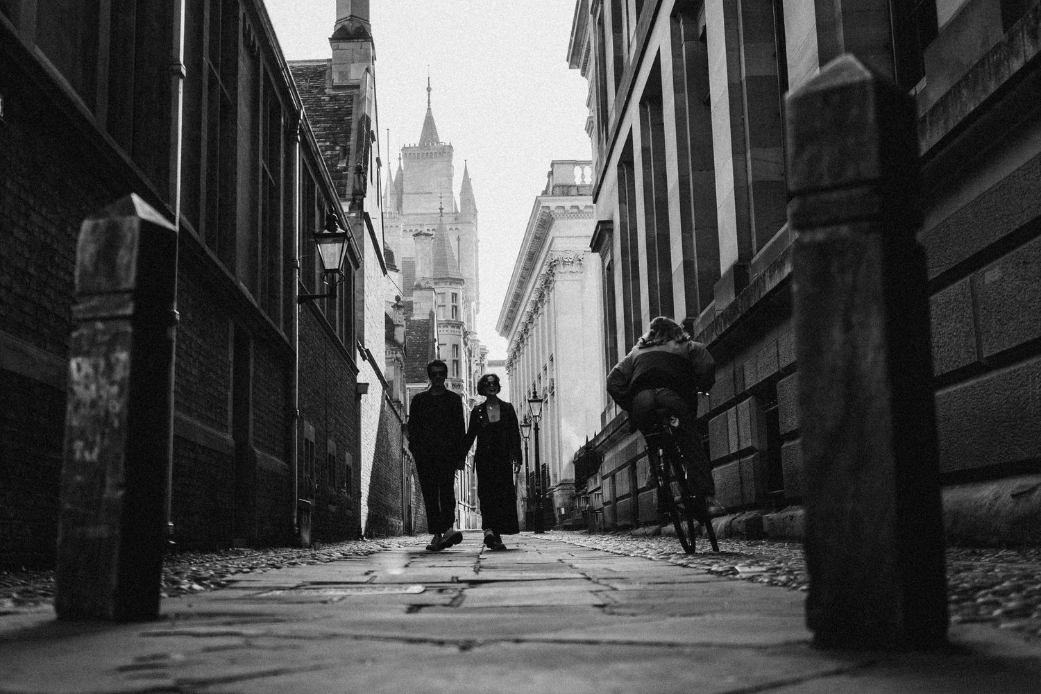 Black and white street scene of pedestrians walking down a narrow alley with church spire visible in background.