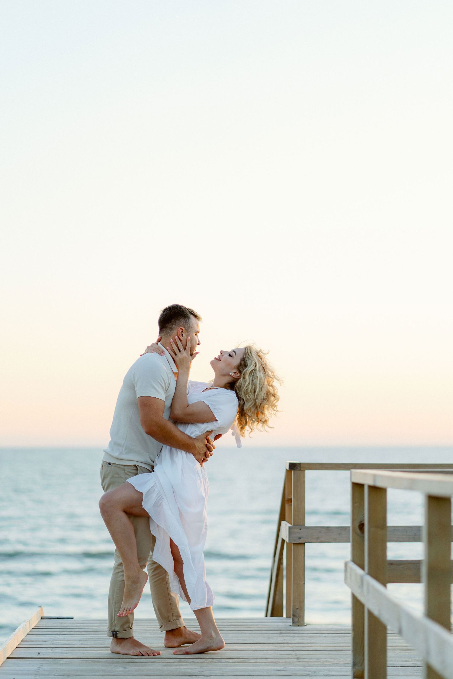 A romantic couple embraces while standing on a wooden dock by the ocean during sunset.