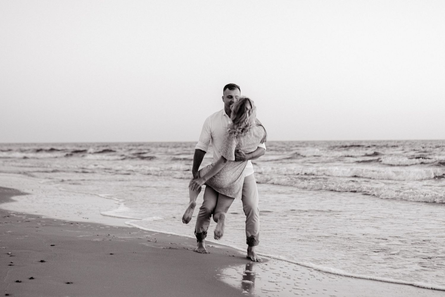 A couple shares a romantic moment while running together on a misty beach in black and white photography.