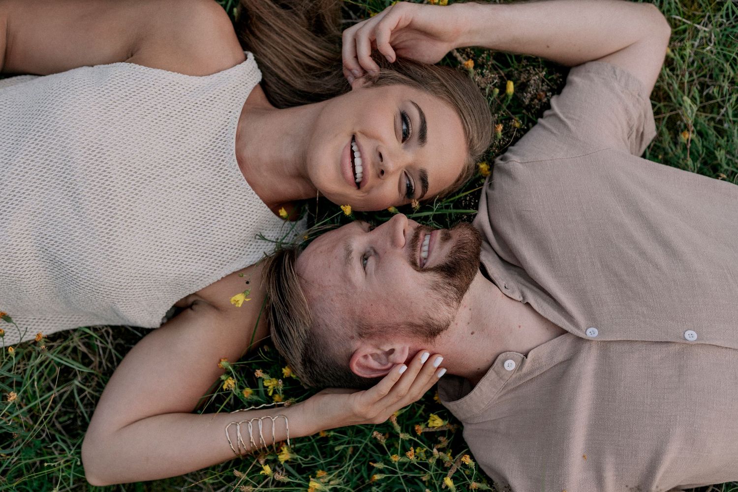 A couple lying together on grass smiling and laughing while wearing tan colored clothing.