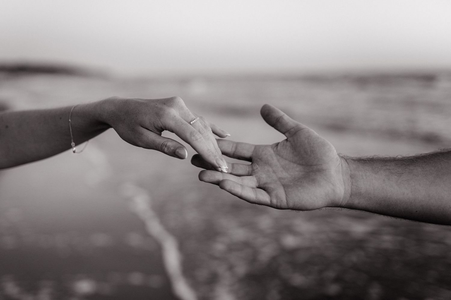 Two hands reaching toward each other with fingertips touching, captured in black and white against a blurred background.