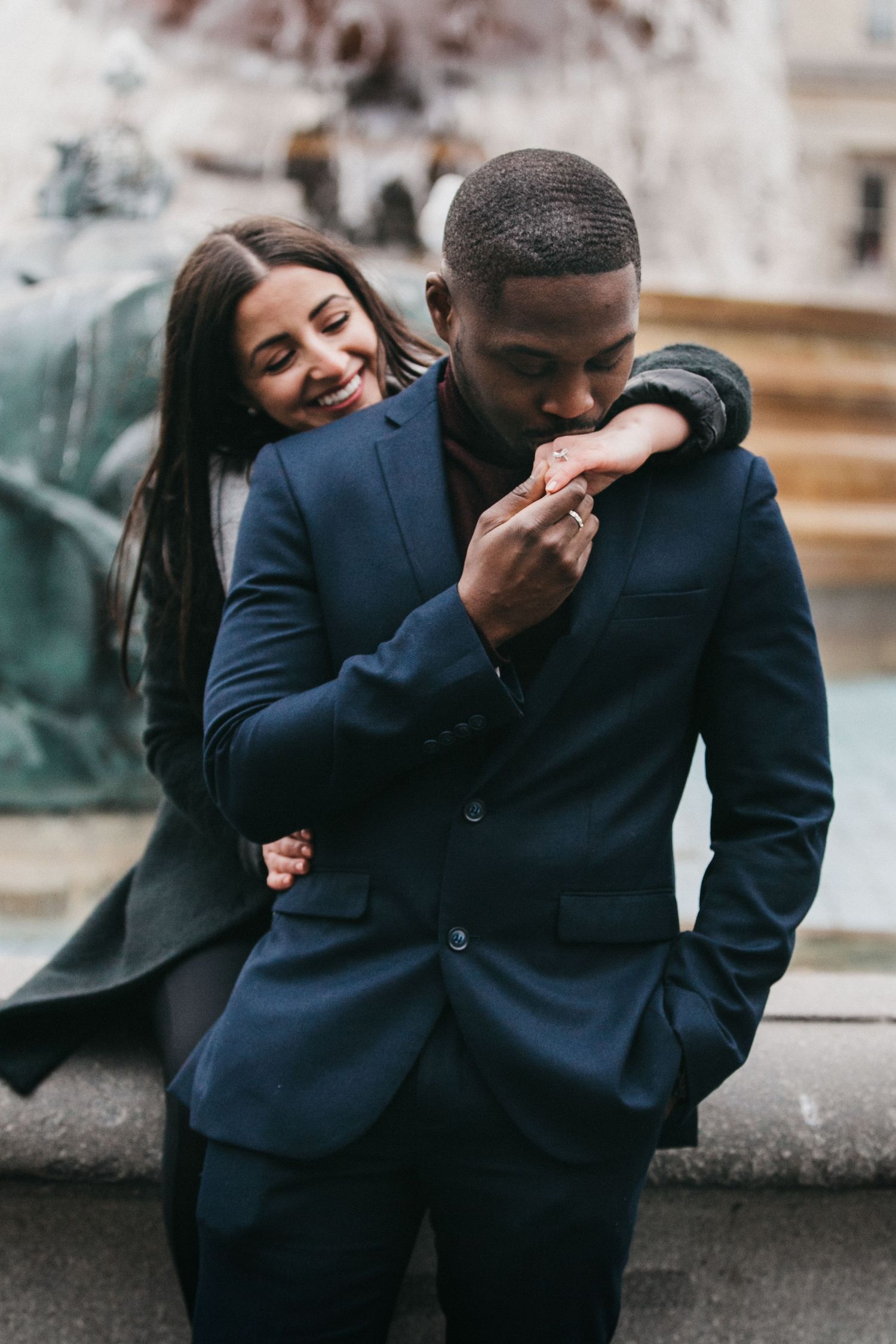 Two people share an intimate moment outdoors, with one embracing the other from behind as they smile and laugh together.