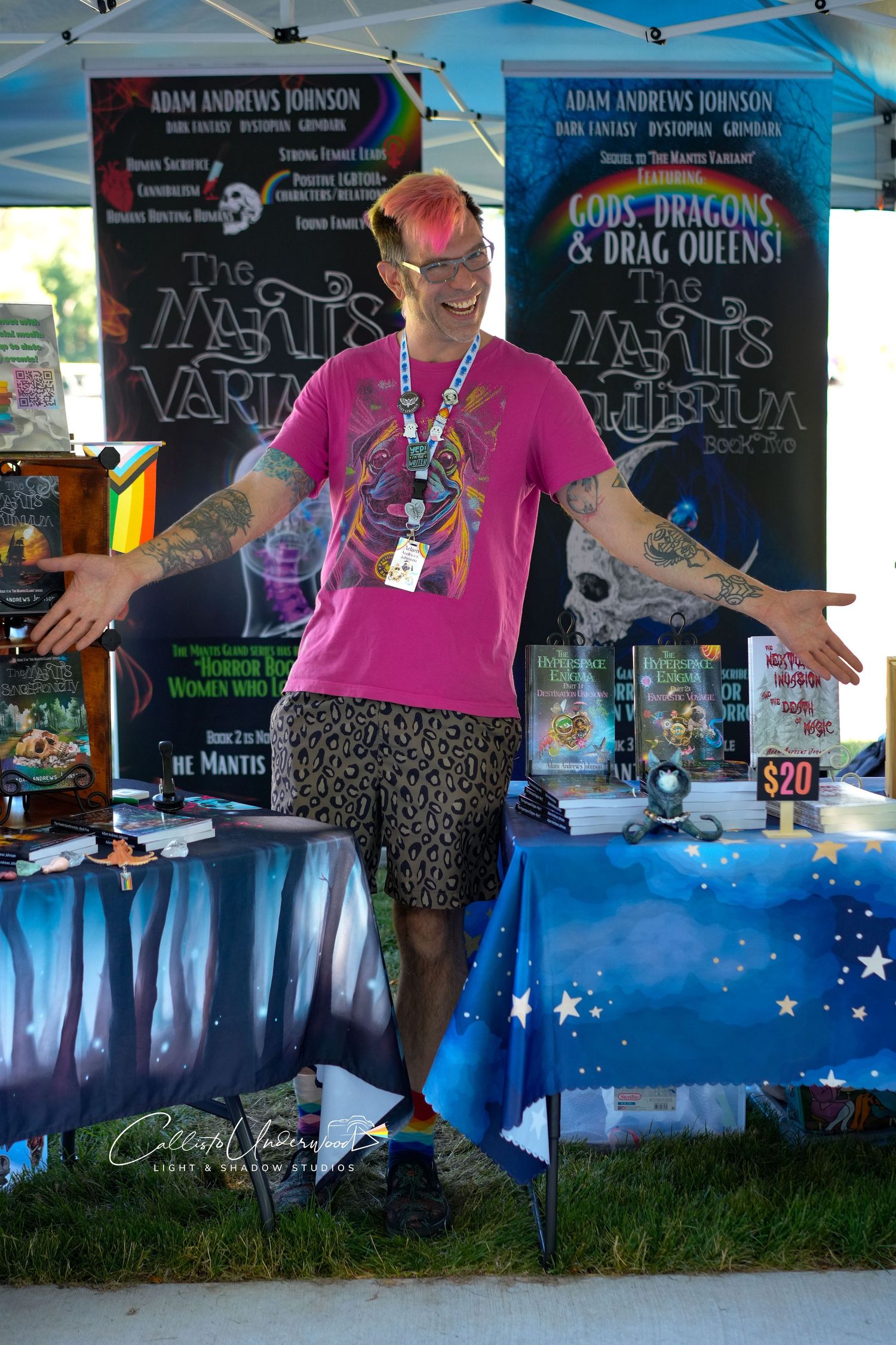 Smiling author with pink-tinted hair and tattoos stands with arms open at an outdoor book booth with fantasy novel banners.