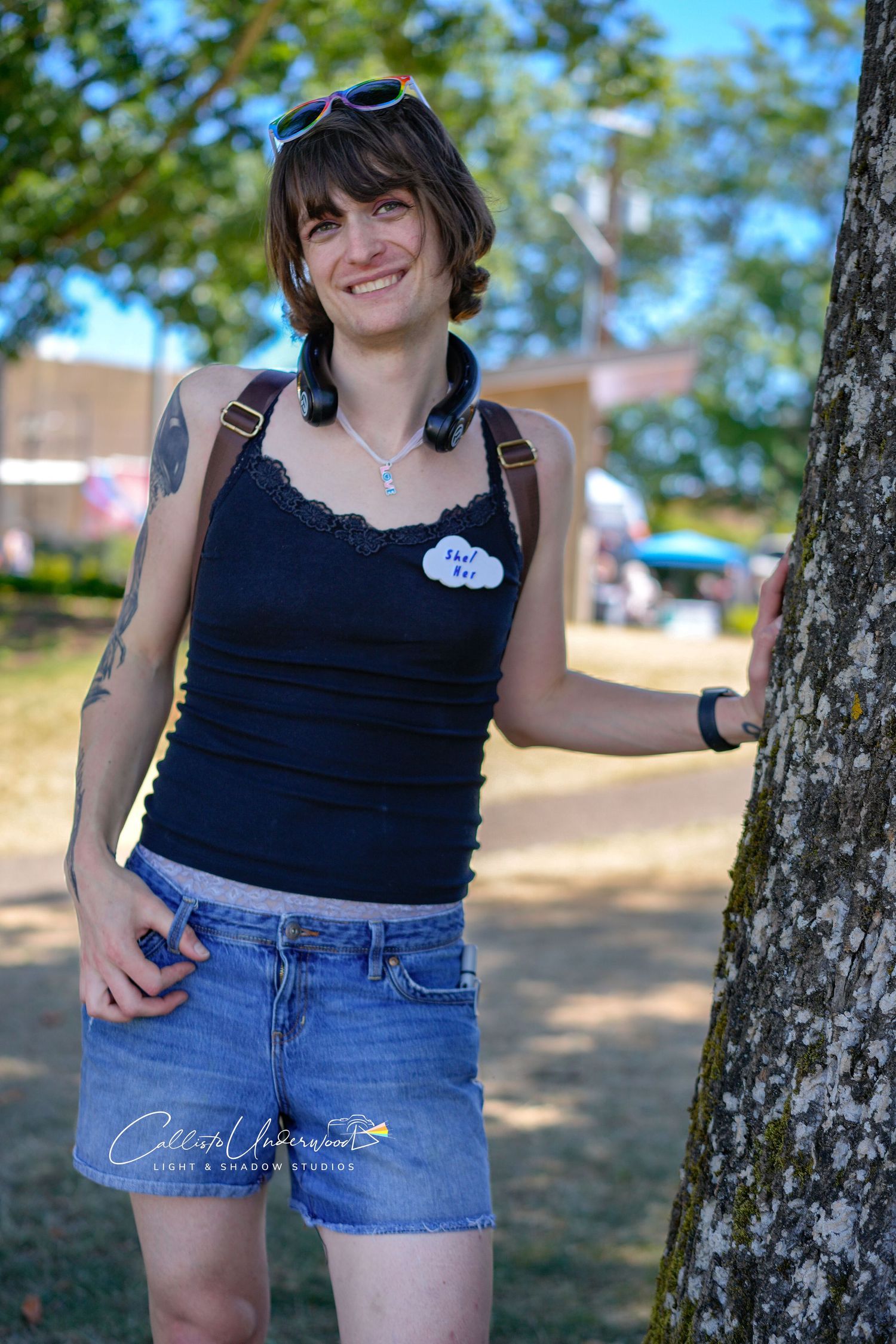 Smiling person in a black lace tank top and denim shorts stands beside a tree at an outdoor park festival.