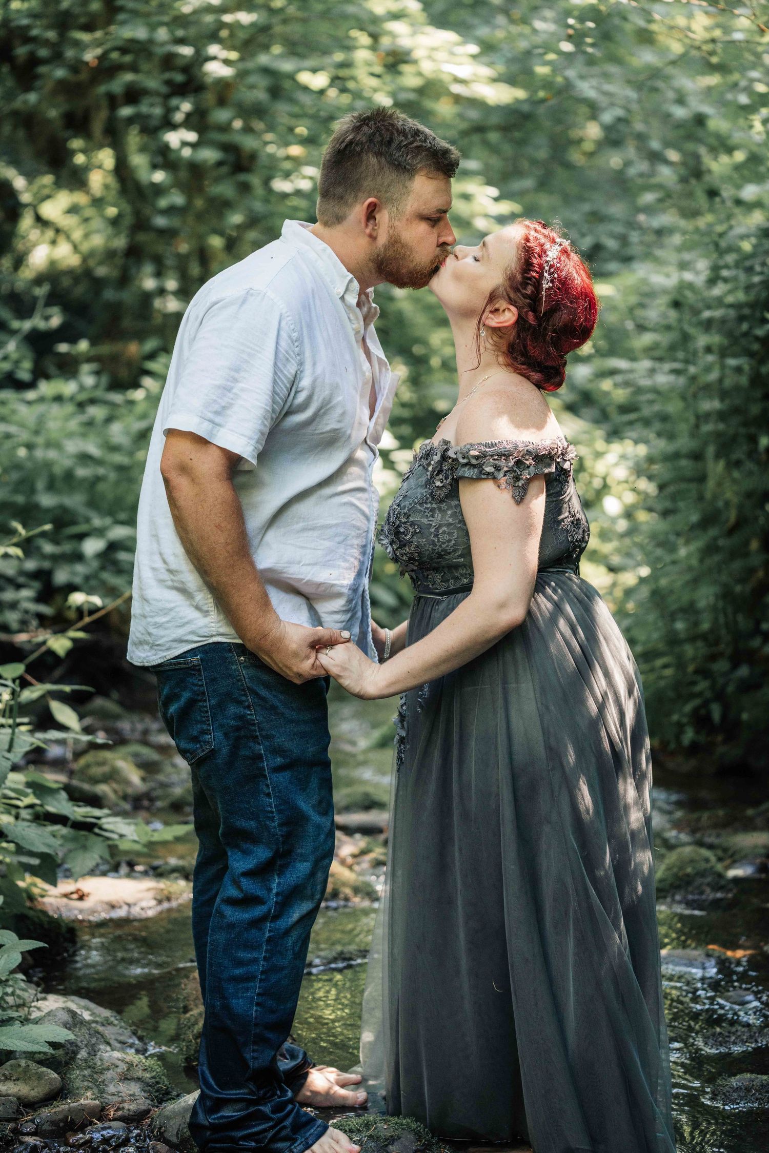 Couple kissing while holding hands in a sunlit woodland creek, the woman wearing a flowing gray off-the-shoulder gown.