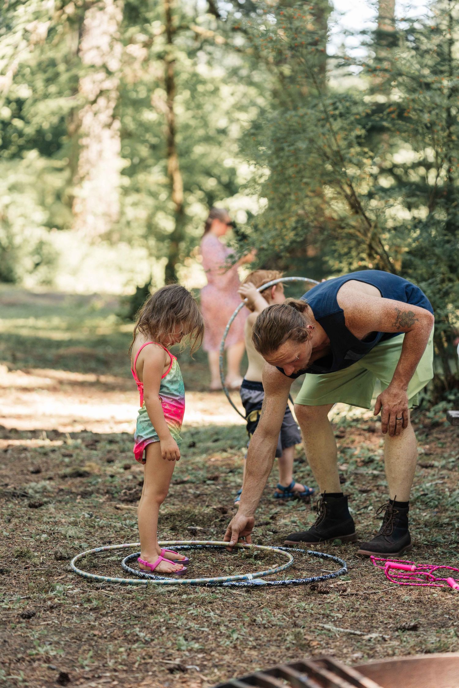 Young girl in a pink swimsuit watches a man in boots arrange hula hoops on the ground at a forest gathering.