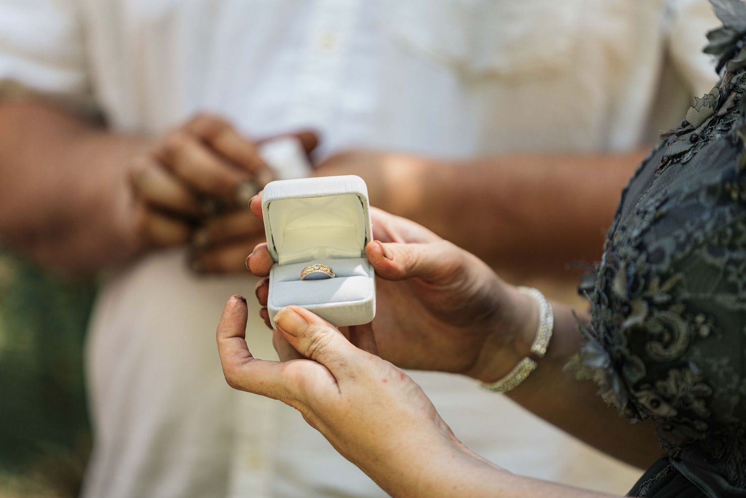 A bride in a lace gray dress holds an open ring box containing a ring toward a man during an outdoor wedding ceremony.