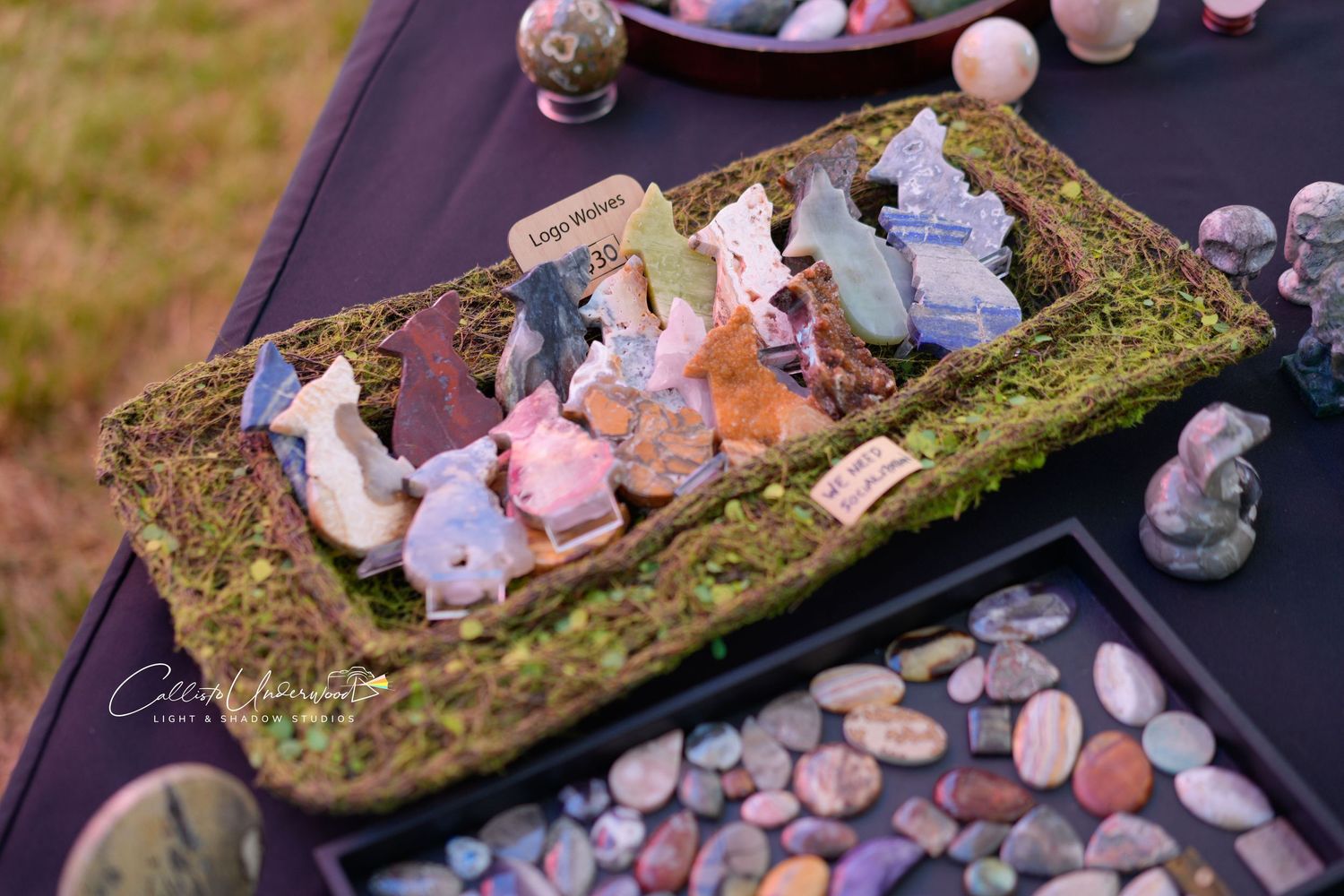 Moss-lined tray of carved crystal animal figurines beside a tray of polished tumbled stones on a vendor table at a market.