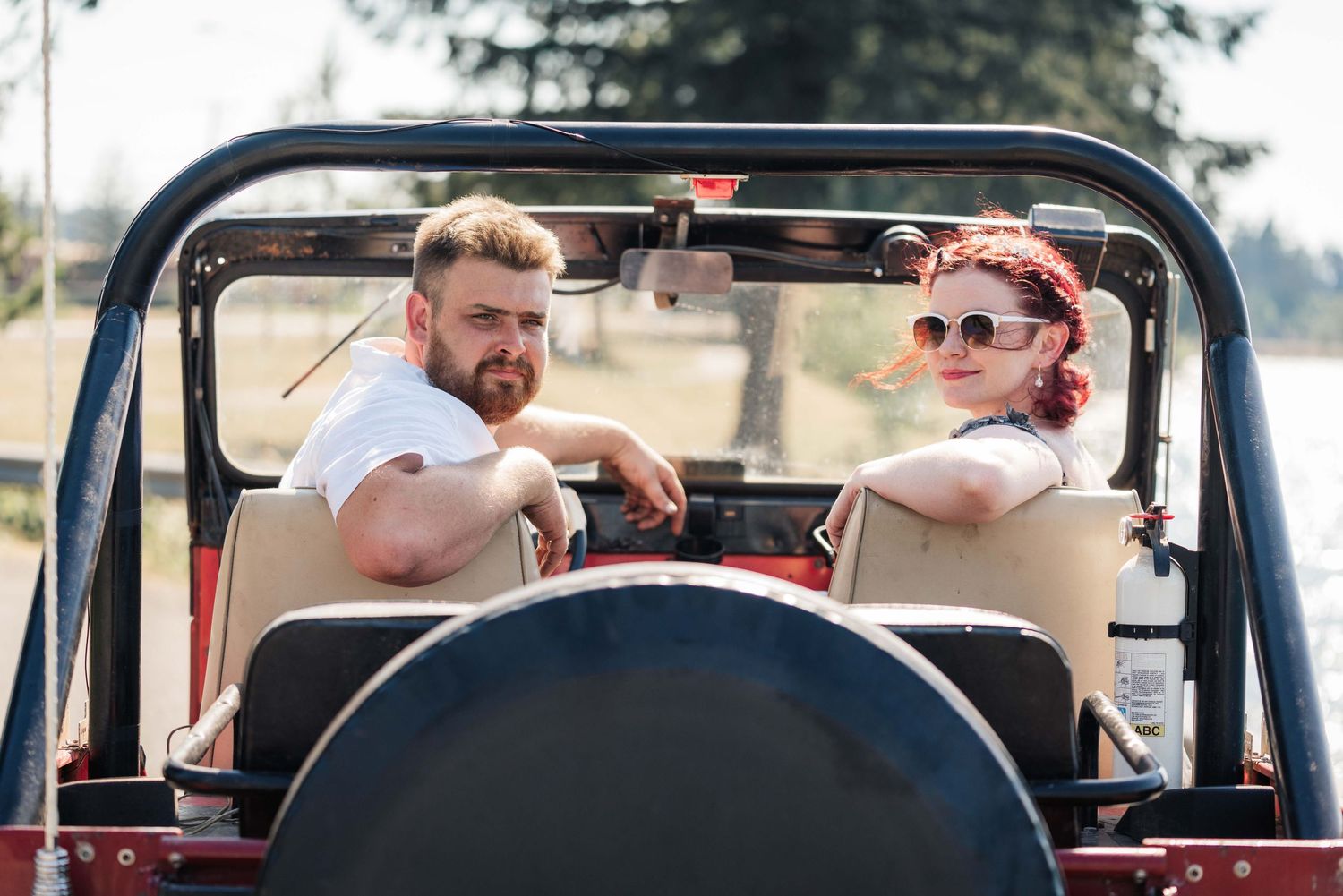 Smiling couple sits in a vintage red open-top Jeep on a sunny day, the woman wearing sunglasses and the man bearded.