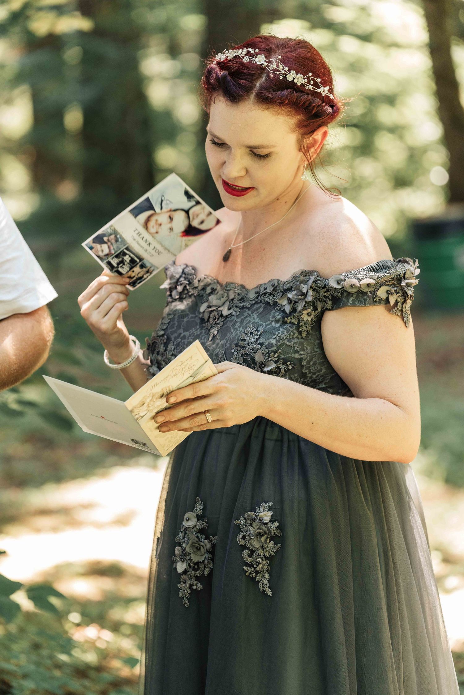 A bride with red hair and a flower crown reads a card and holds a small photo booklet during an outdoor forest wedding ceremony.