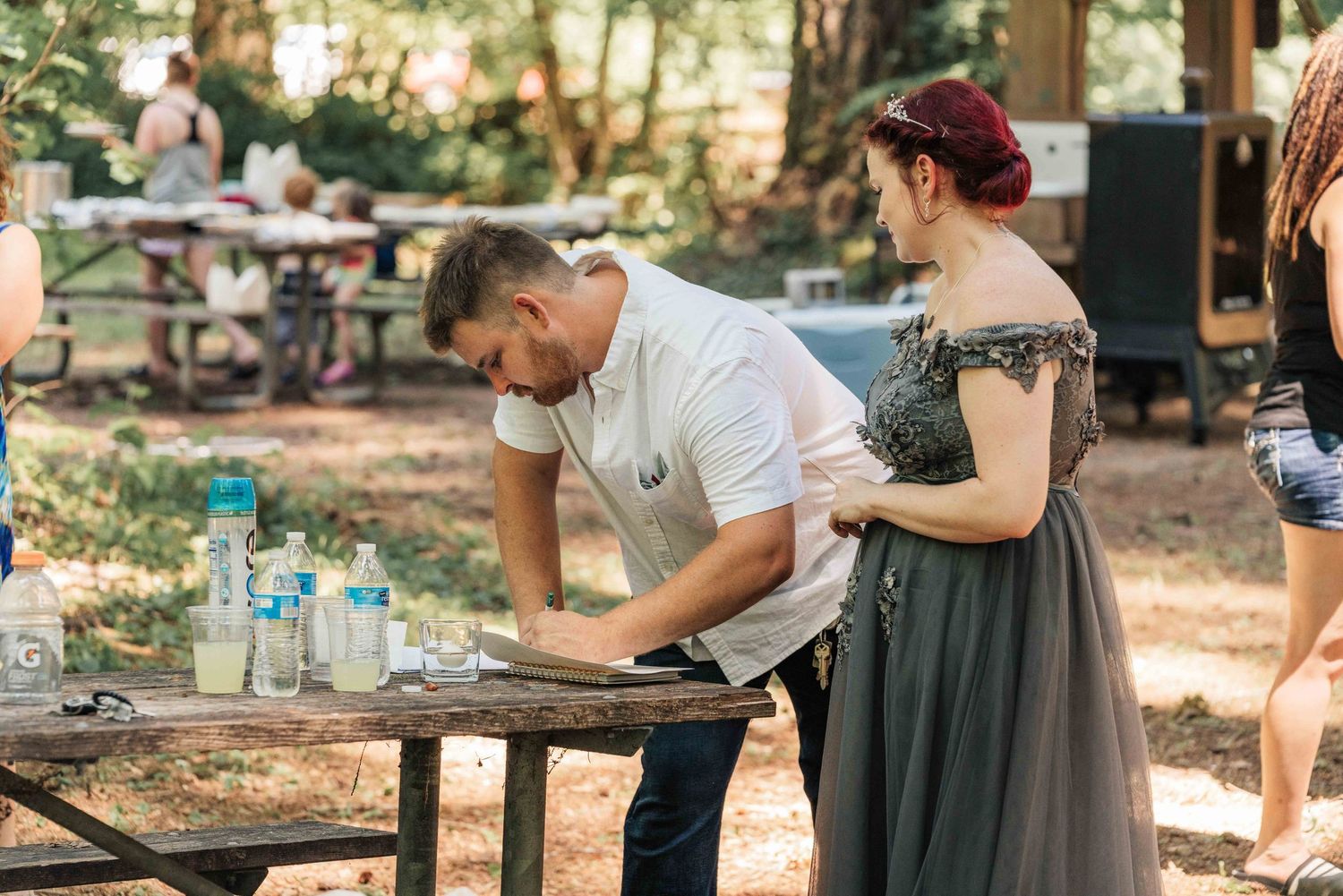 Man in a white shirt signs a document at a wooden picnic table beside a woman in a gray gown at an outdoor forest event.