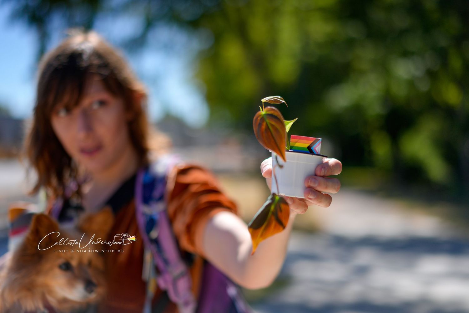 Woman in an orange shirt holds a small cup with a colorful flower toward the camera at an outdoor event with blurred trees behind.