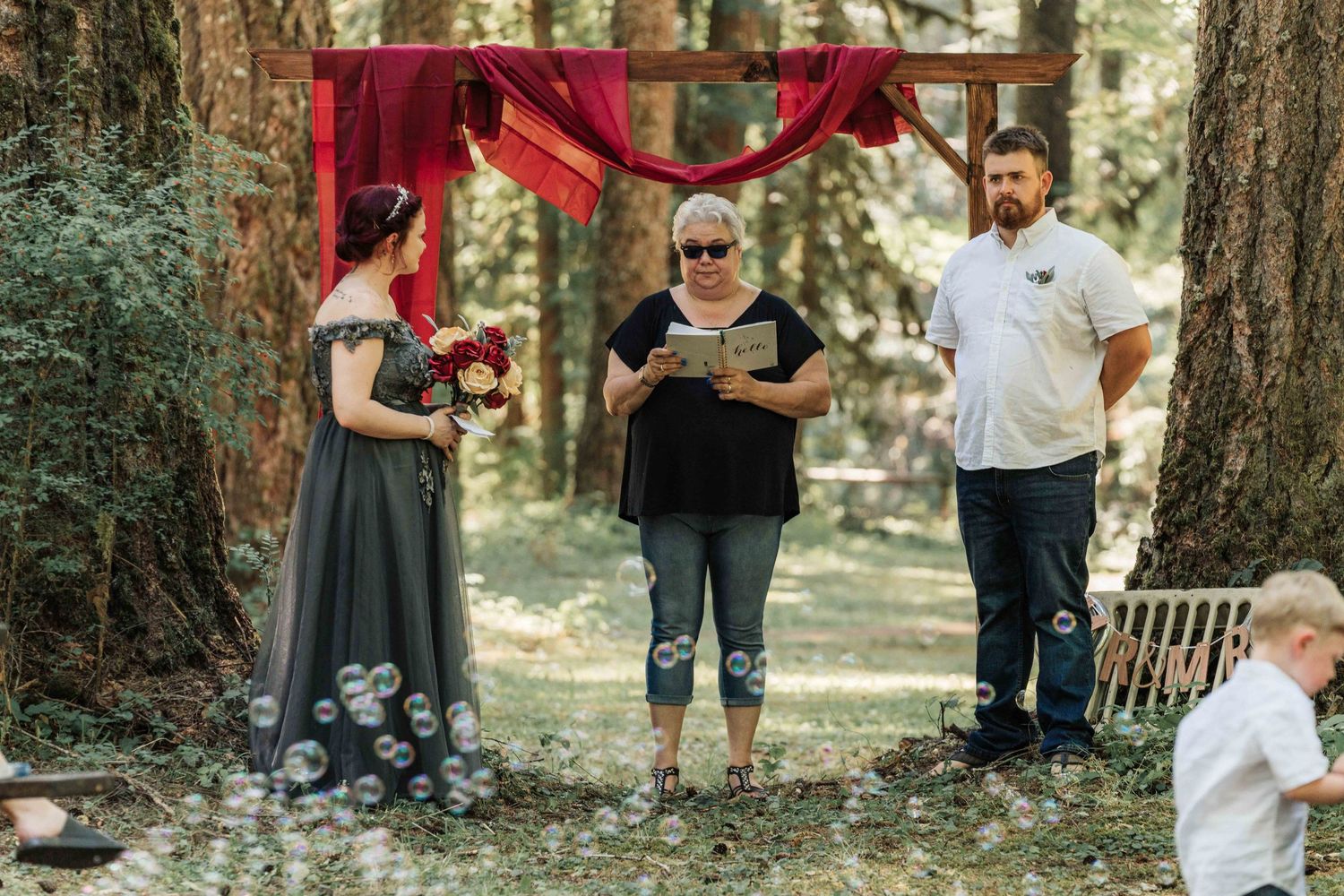 A bride holds a red bouquet while the officiant reads and the groom watches, with a toddler in the foreground among bubbles.