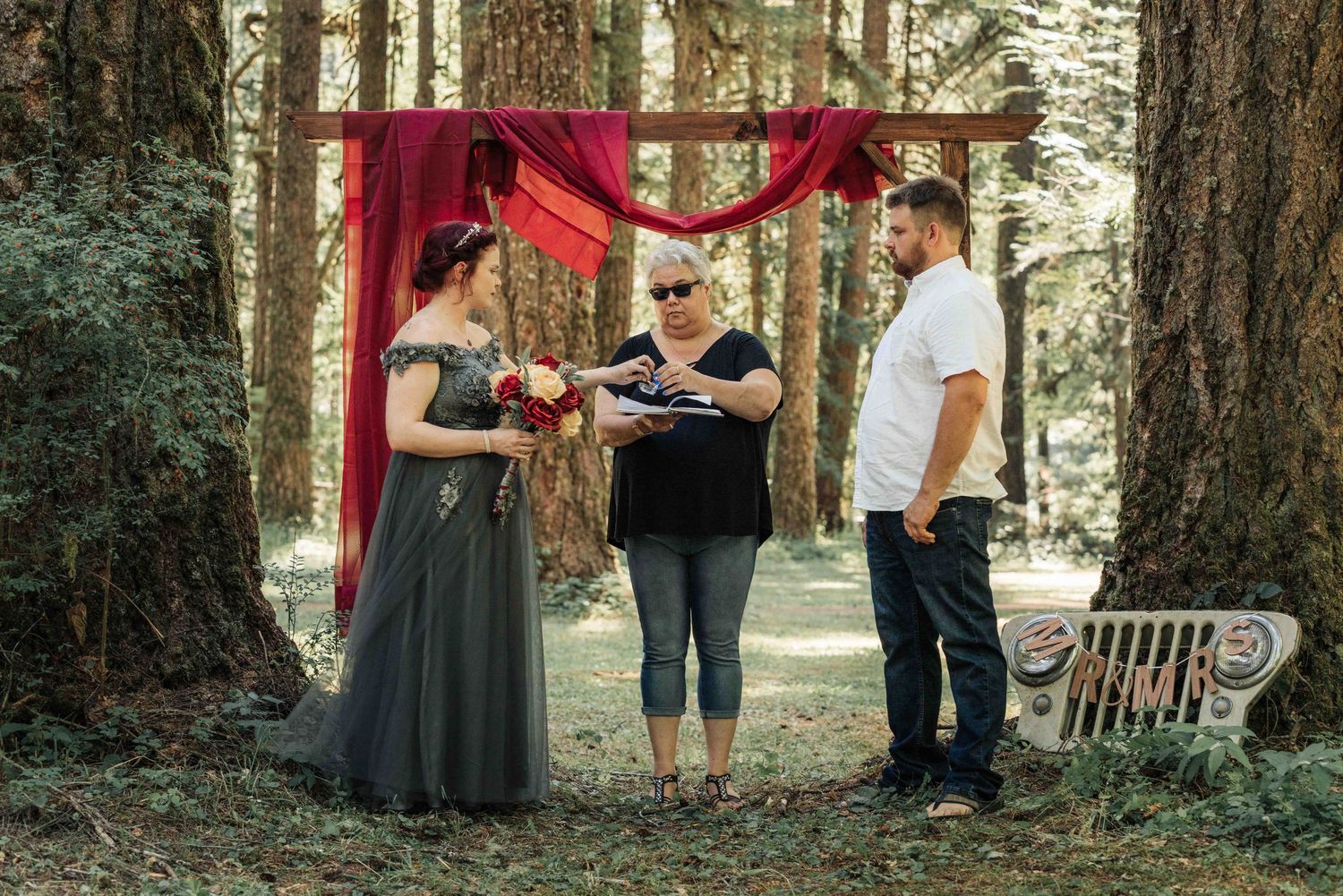 A bride in a gray gown and a groom in a white shirt exchange vows with an officiant under a red draped arch in a forest.