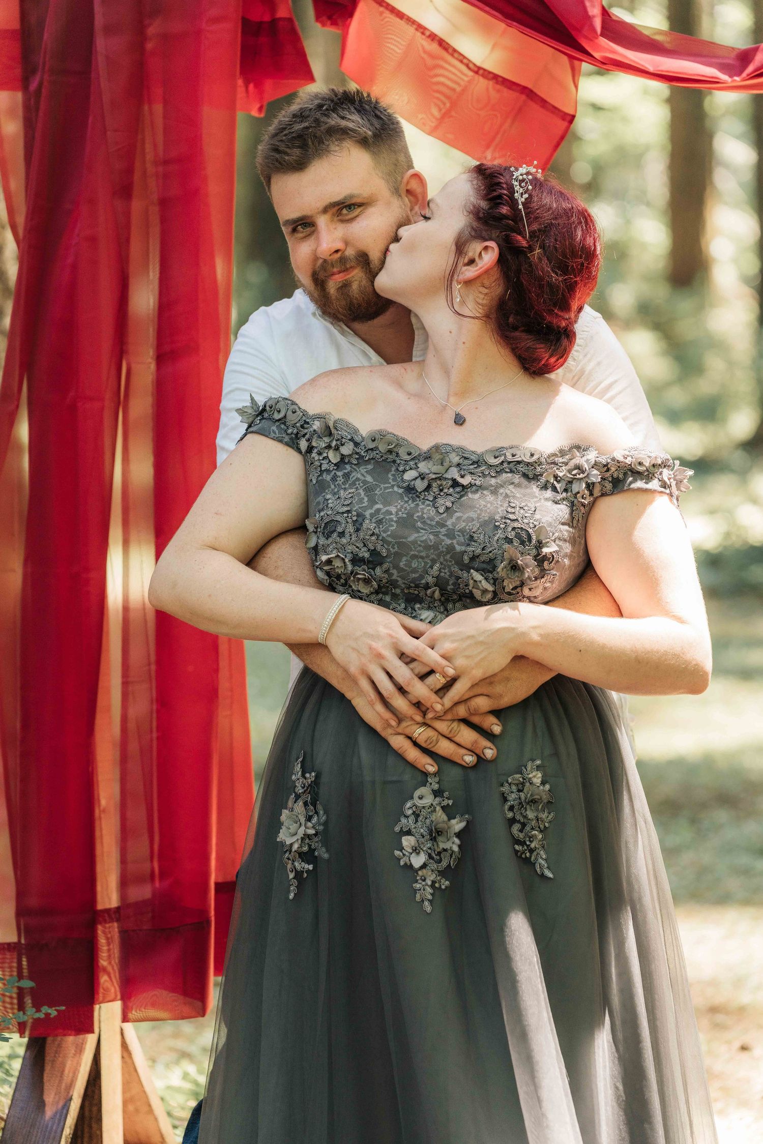 Bearded man in a white shirt embraces a red-haired woman in a gray embroidered gown from behind beneath red drapes in the forest.