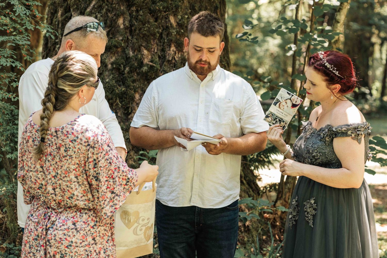 A bearded groom and a red-haired bride in a gray gown stand with an officiant and a braided-hair woman during a forest wedding ceremony.