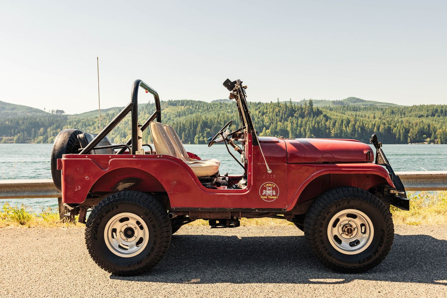 Side view of a restored vintage red Jeep parked along a lakeside road with evergreen-covered hills behind it.