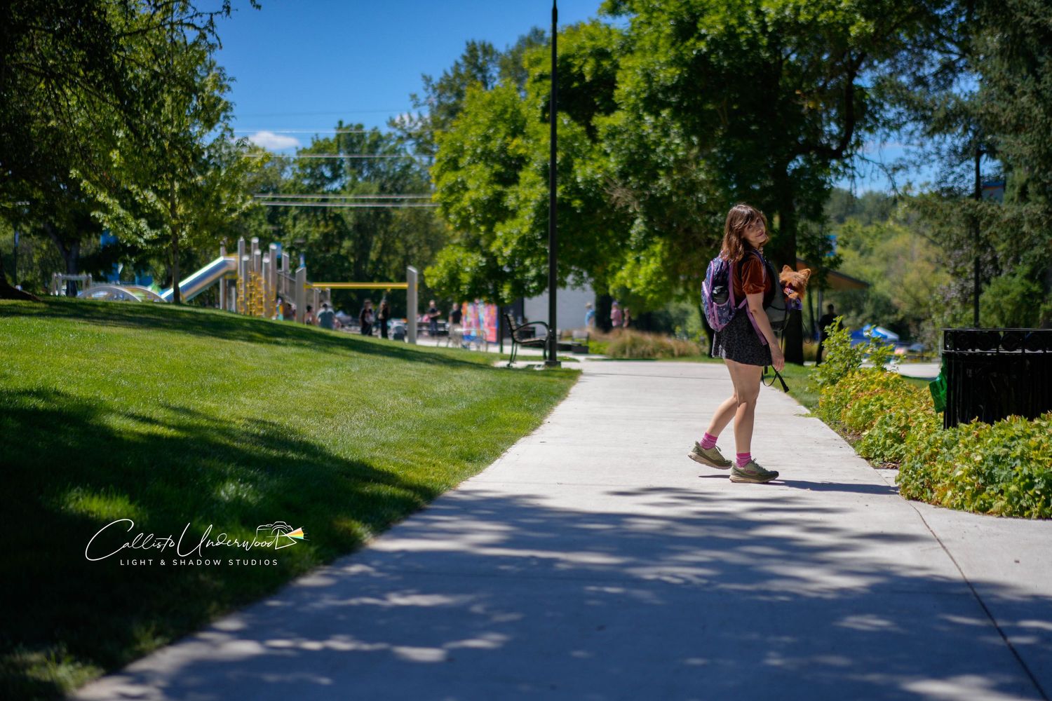 Young woman with a backpack walks along a sunlit paved path beside a green lawn and trees in a park with a playground in the distance.